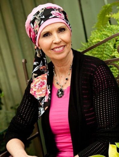 Woman with pink and black floral head covering smiles outdoors, wearing pink shirt and black cardigan.