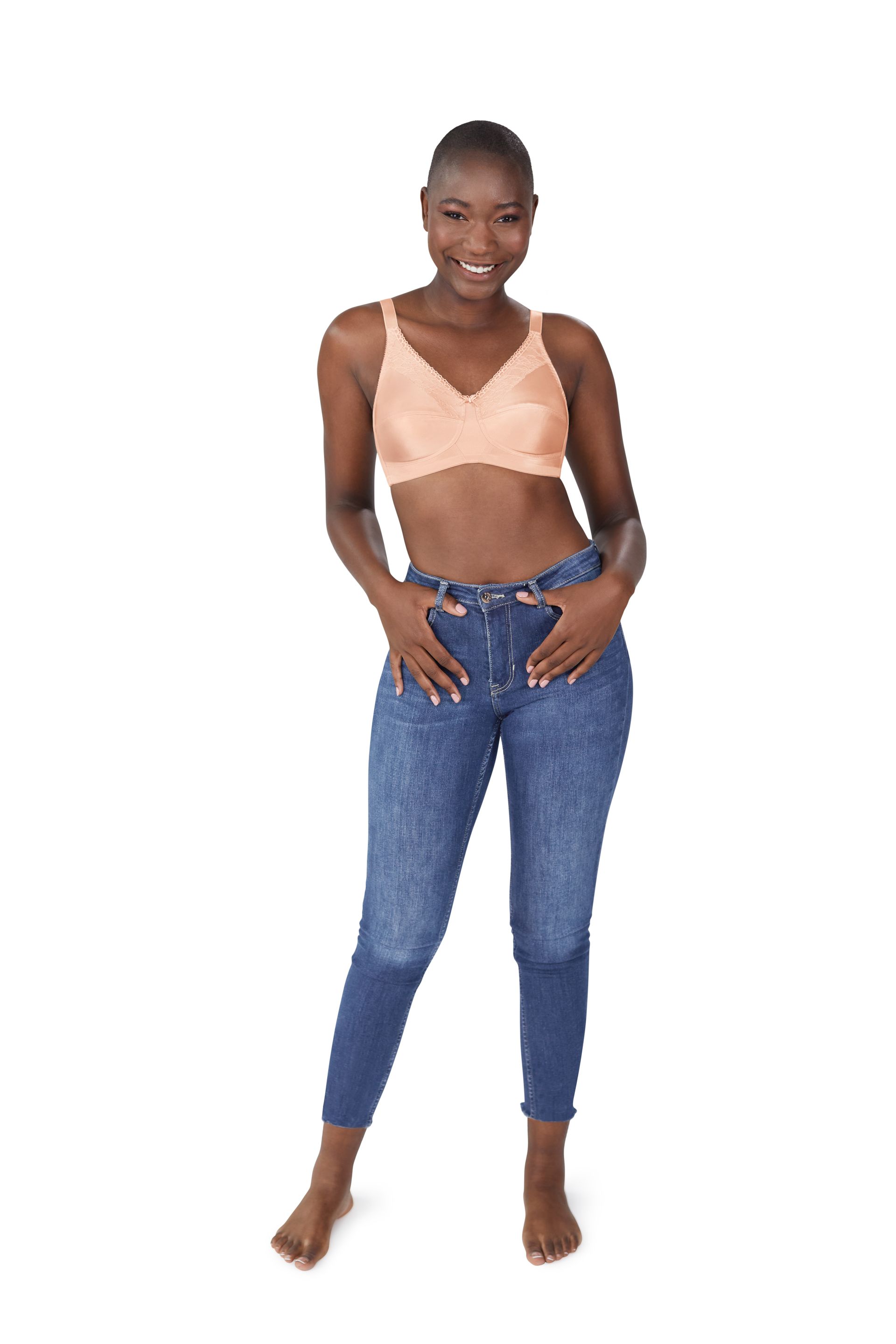 Woman in a light peach-colored bra and blue jeans smiles, with hands in pockets; white background.