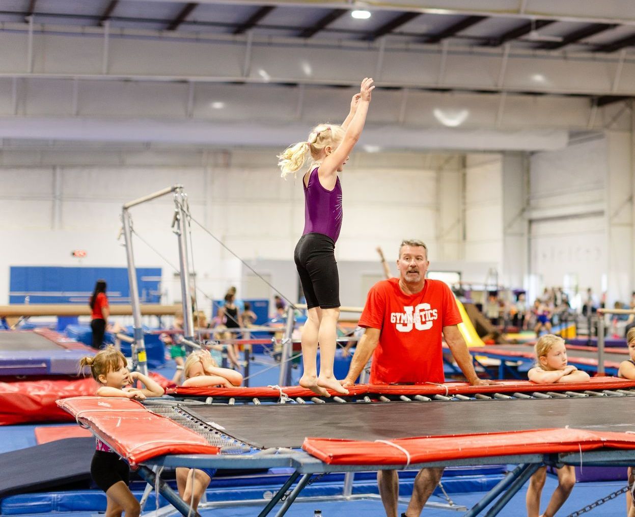 A girl is jumping on a trampoline in a gym while a man watches.