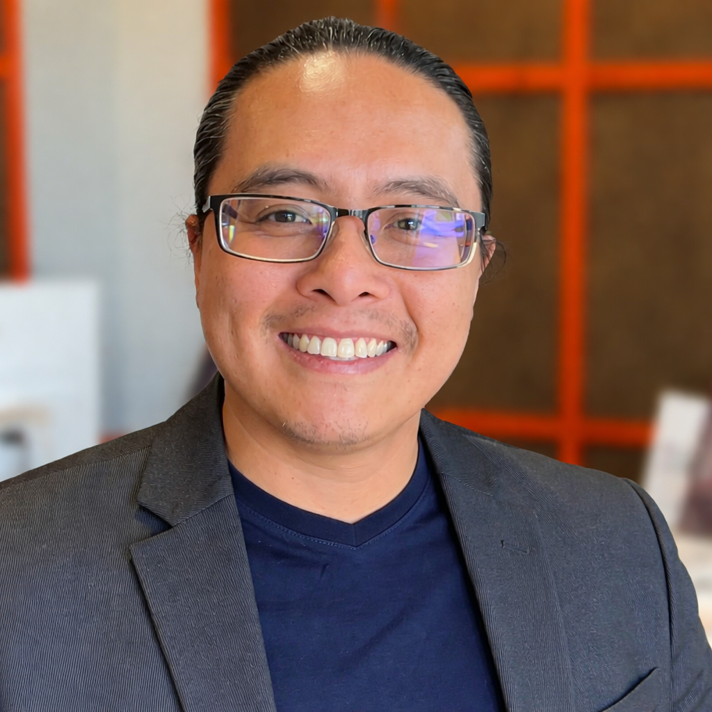 Man with glasses smiles, wearing a navy shirt and gray blazer in a bright office setting.