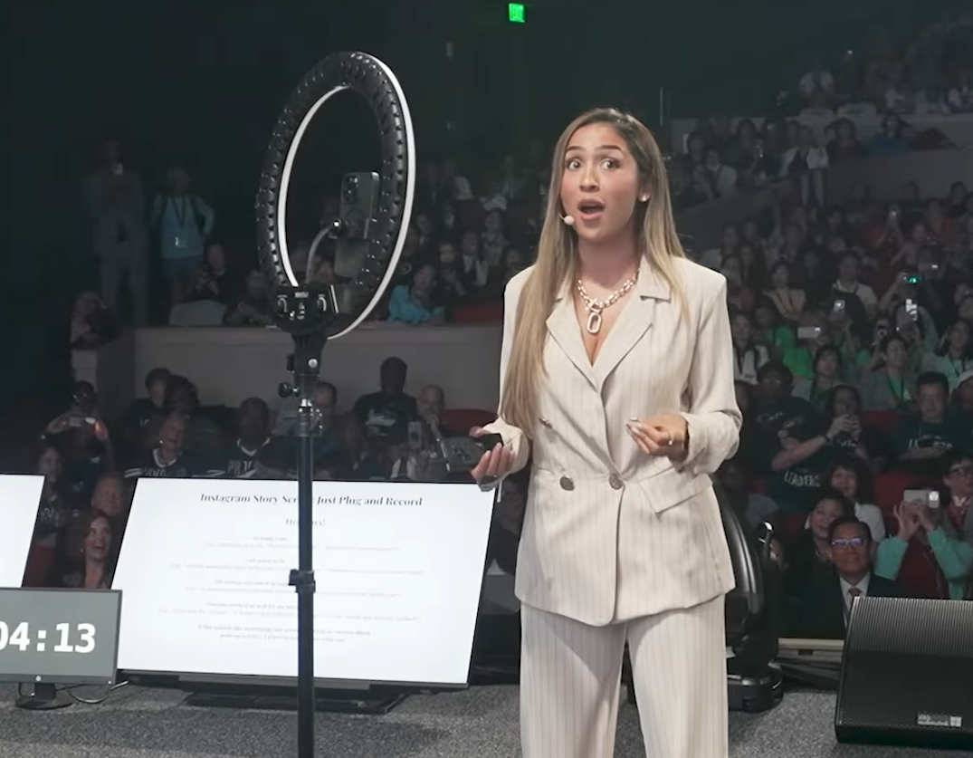 Woman in beige suit speaks at conference, gesturing, with ring light and crowd in background.