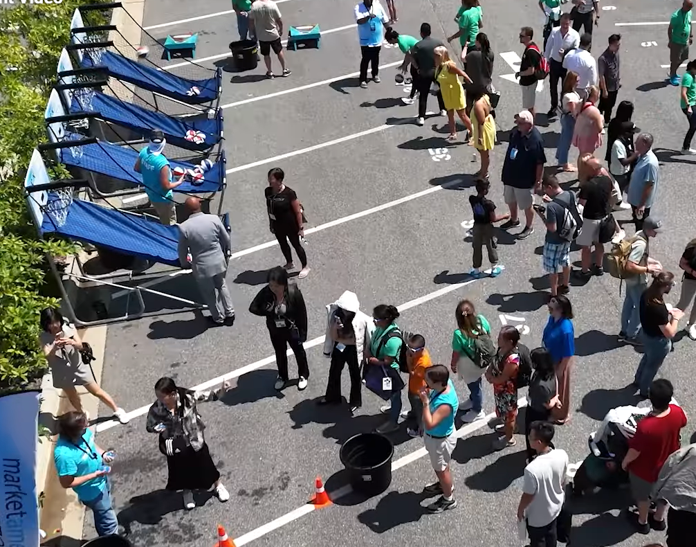 People gathered outdoors at an event, near tents and a parking lot.