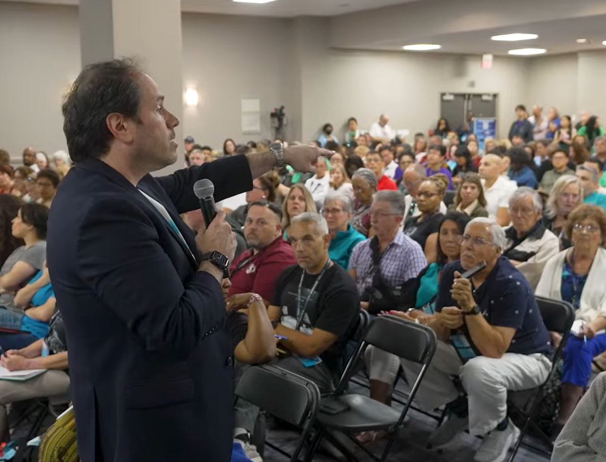 Man speaking to a large audience, pointing. Attendees seated in rows, some wearing name tags, in a large room.