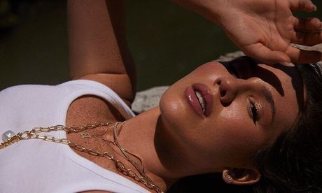 Woman with golden necklace and sunkissed skin lies down, hand shading face.