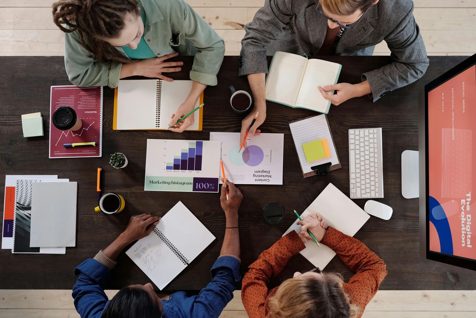Four people collaborating at a table, reviewing documents and writing in notebooks. Charts and keyboard on the table.