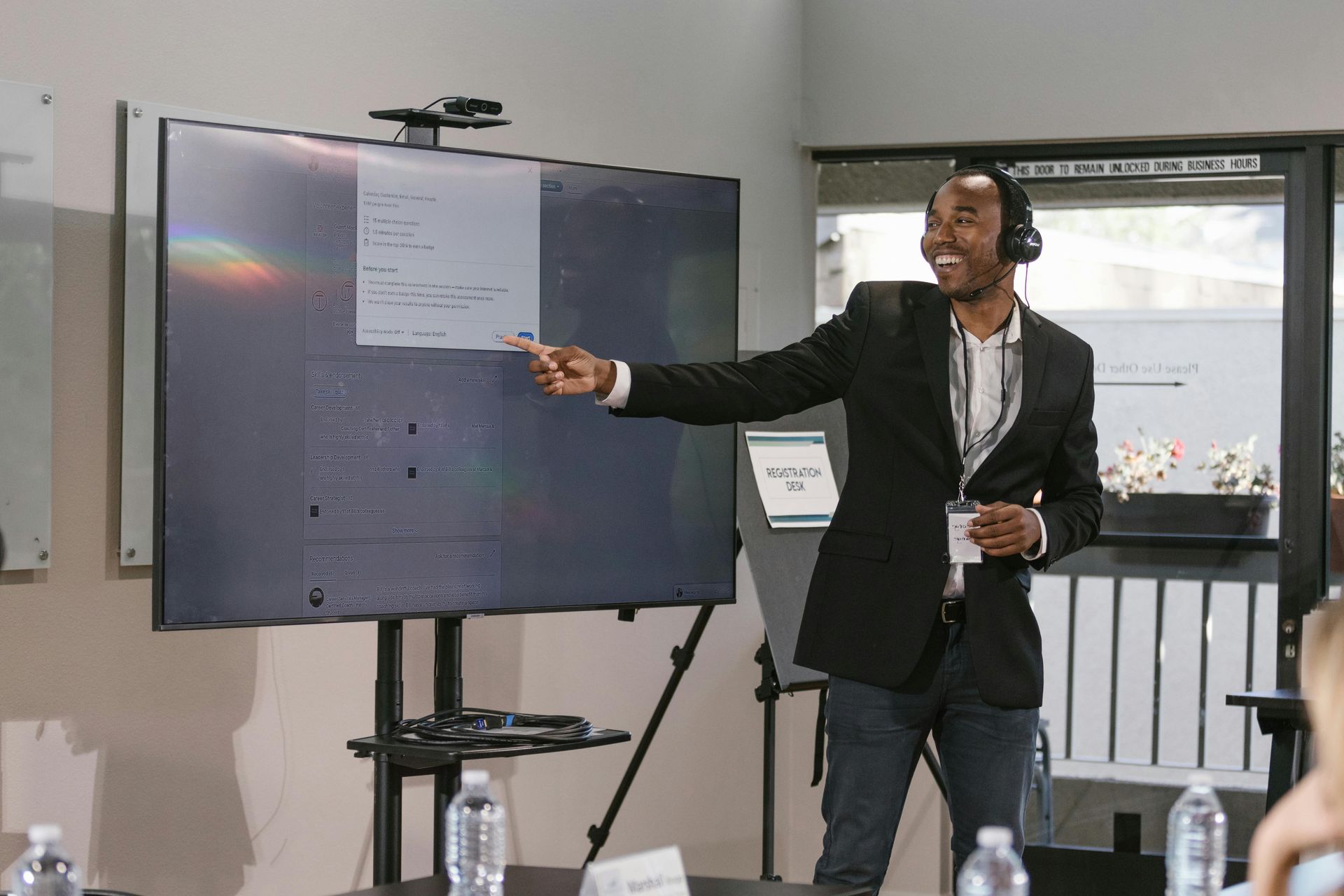 Man in suit points at a screen displaying code, presenting in a bright room.