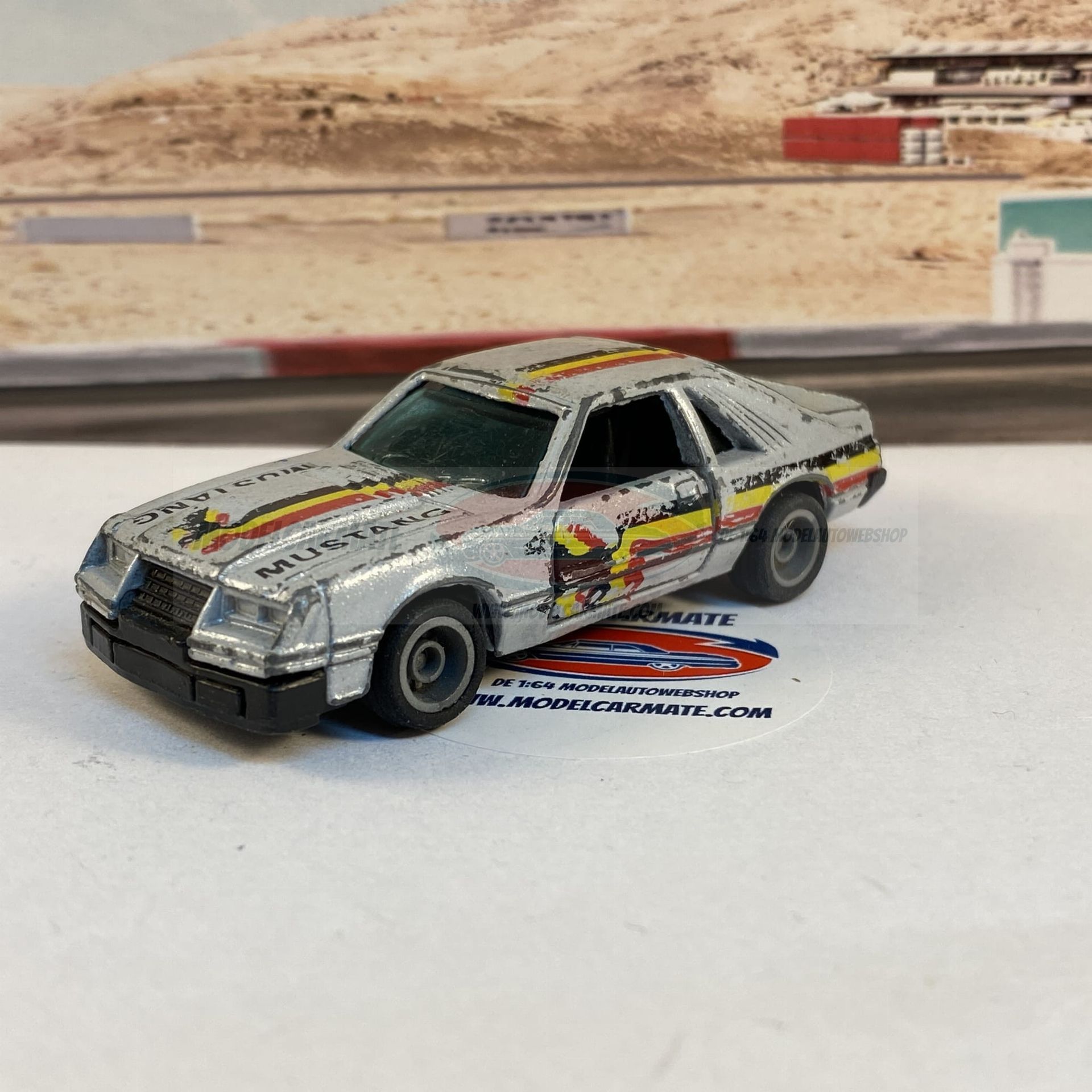 Zilverkleurige speelgoed-Ford Mustang met racestrepen op een racebaanachtergrond.