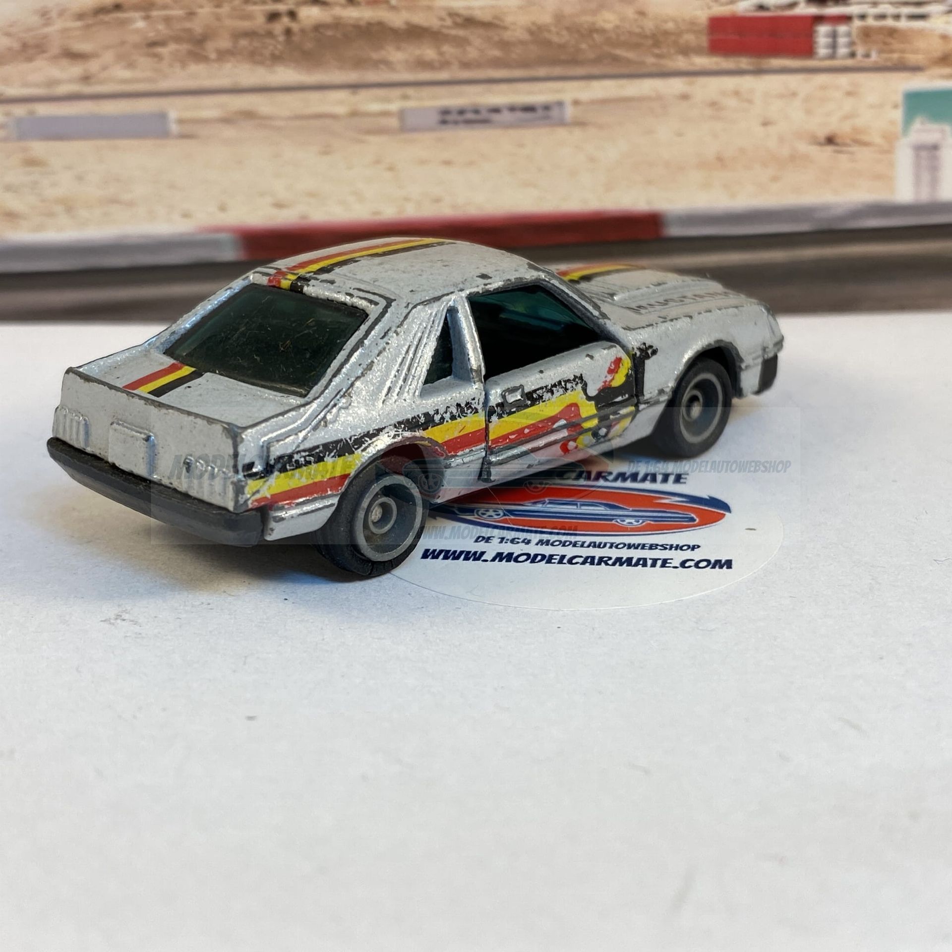 Zilverkleurige speelgoed-Ford Mustang met racestrepen op een racebaanachtergrond.