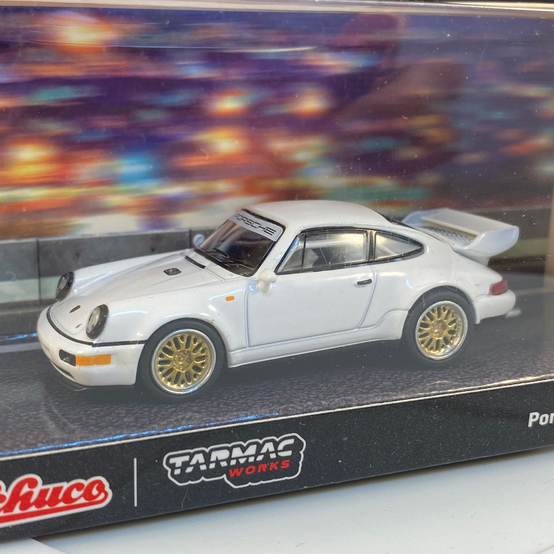 Witte Porsche 911 modelauto met gouden velgen, in een vitrine met een achtergrond van een nachtelijk straatbeeld.