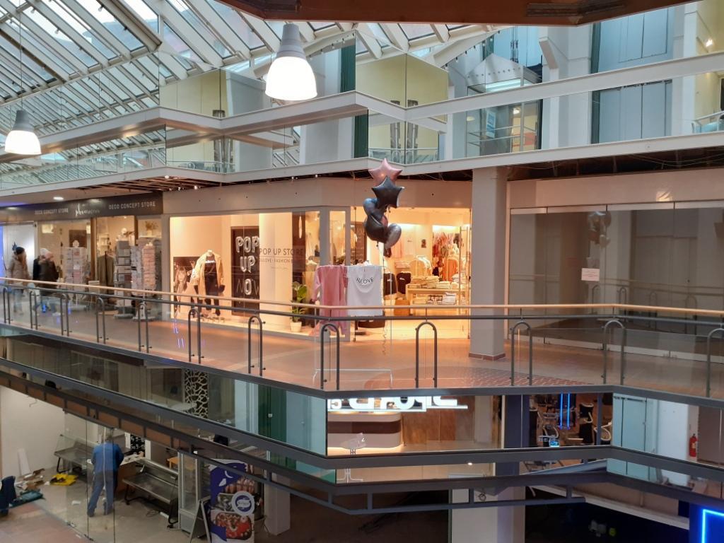 Innenansicht eines mehrstöckigen Einkaufszentrums mit Glasgeländern, Oberlichtern und hängenden Skulpturen.