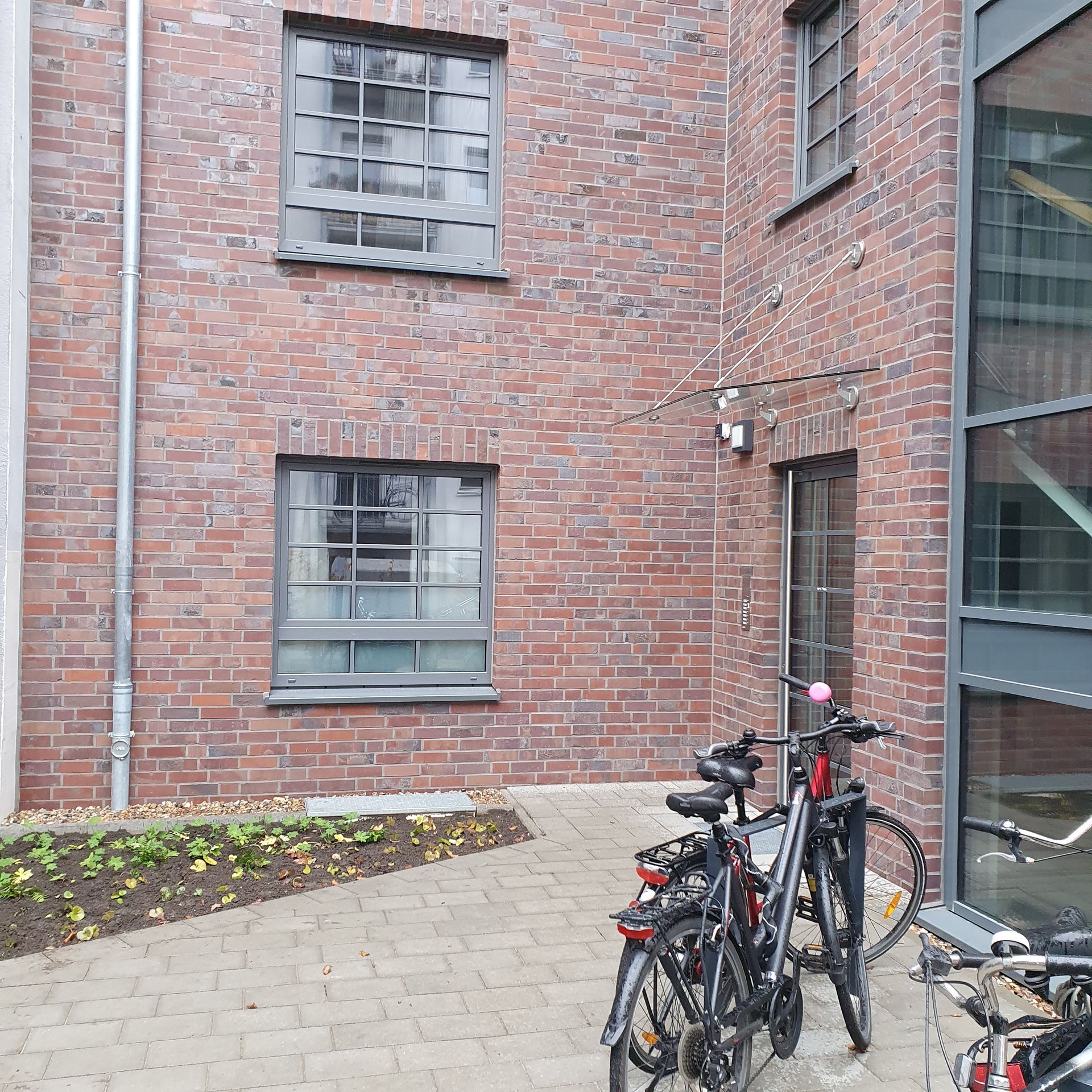 Ein mit Ziegeln gepflasterter Innenhof neben einem Gebäude, in der Nähe einer Glastür sind mehrere Fahrräder geparkt.