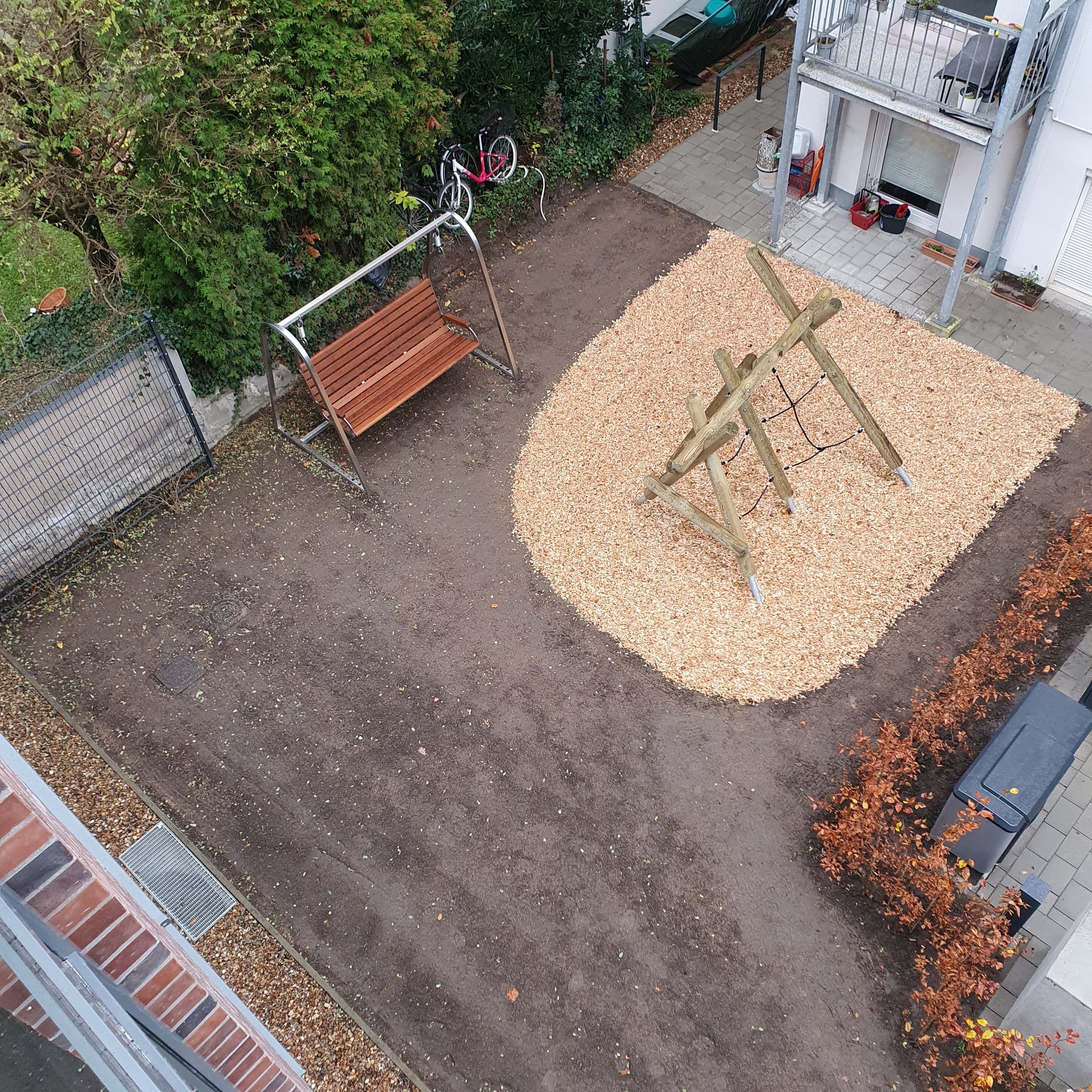 Kleiner Spielplatzhof mit einer hölzernen Schaukelbank und einer Wippe auf Kies neben einem Gebäude.