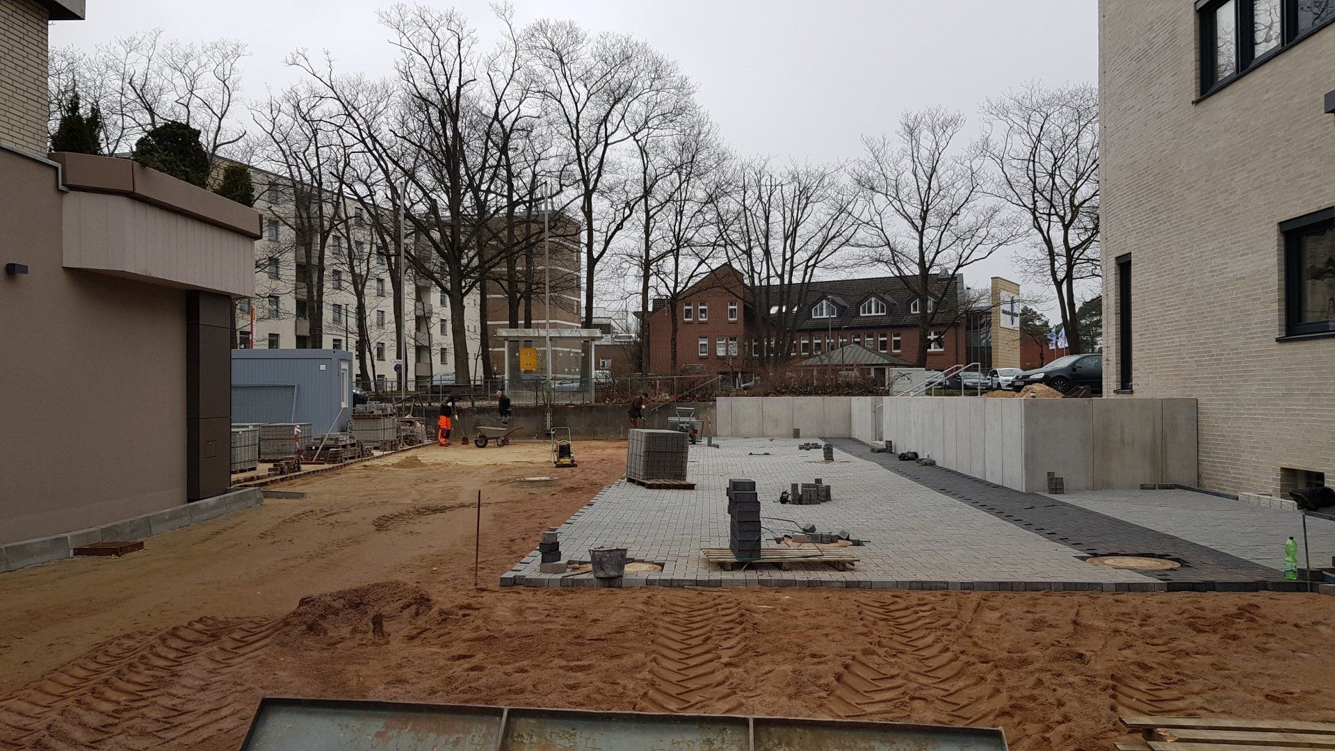 Baustelle mit Kieswegen, Betonmauern und kahlen Bäumen unter einem bedeckten Himmel