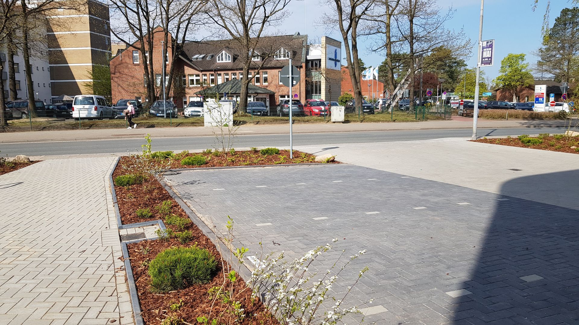 Ruhiger Stadtplatz mit gemauerten Pflanzbeeten, gepflastertem Gehweg und Straße mit Bäumen und Gebäuden im Hintergrund