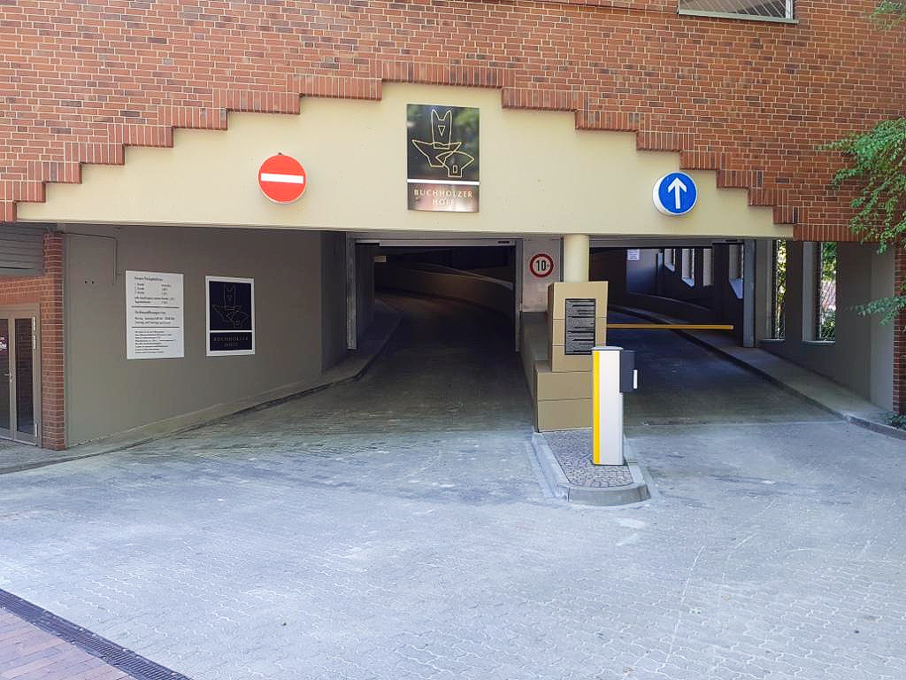 Einfahrt zur Parkgarage mit niedrigen Schildern, Backsteinfassade und dunkler Durchfahrt.
