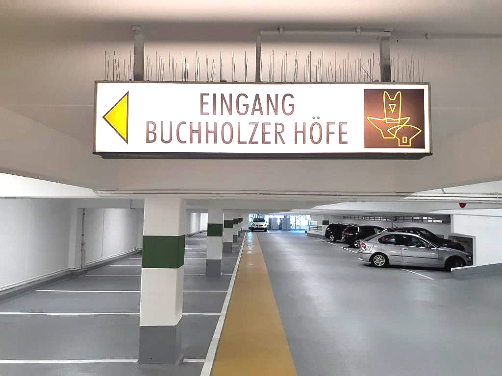 Eingangsschild der Parkgarage mit der Aufschrift „Eingang Buchholzer Höfe“, im Hintergrund parkende Autos.