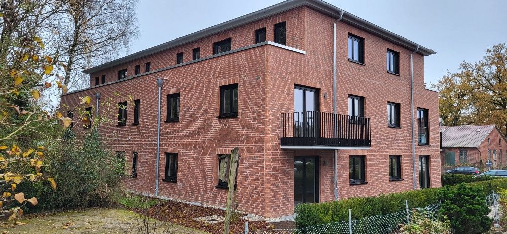 Dreistöckiges Wohnhaus aus roten Backsteinen mit schwarzen Balkonen, umgeben von Bäumen und Sträuchern.