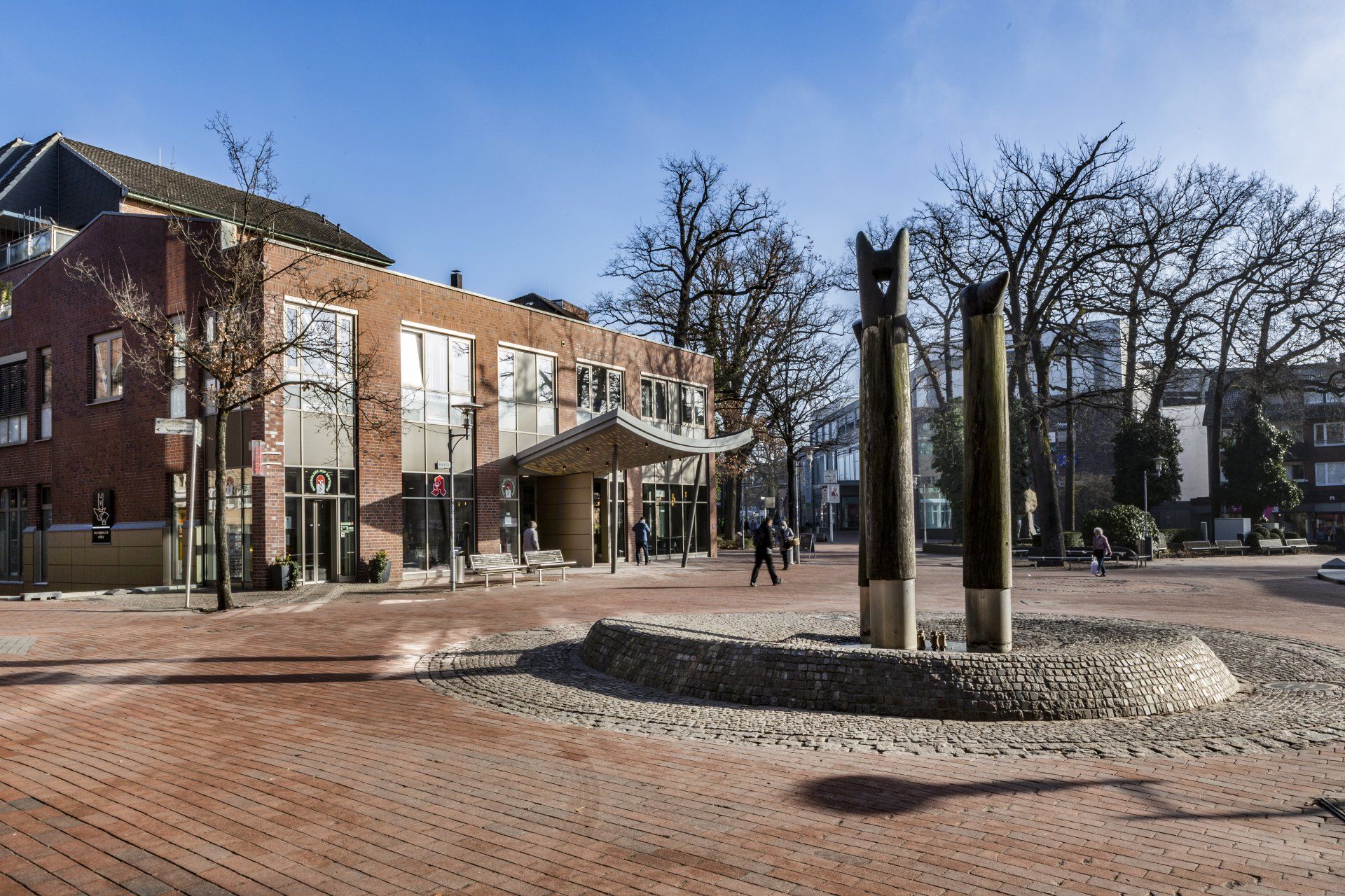 Ein Backsteinplatz mit einem Backsteingebäude, kahlen Bäumen und einer zentralen Skulptur unter klarem Himmel.