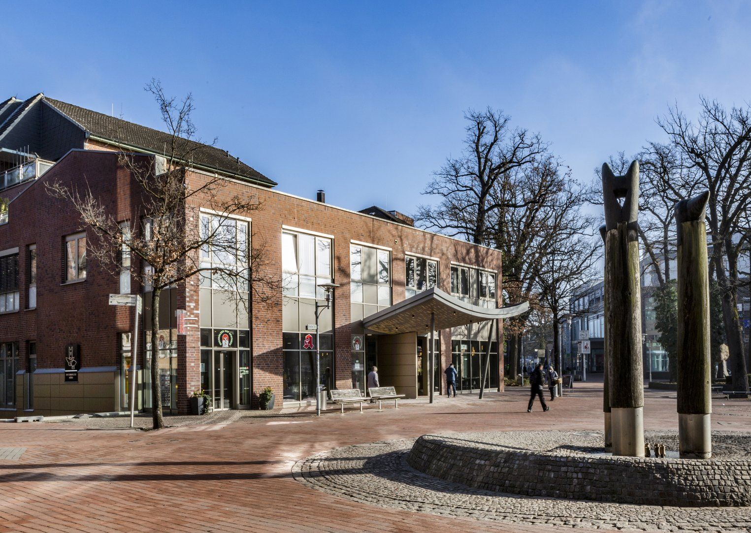 Modernes Backsteingebäude mit großen Fenstern und überdachtem Eingang auf einem Platz an einem sonnigen Tag
