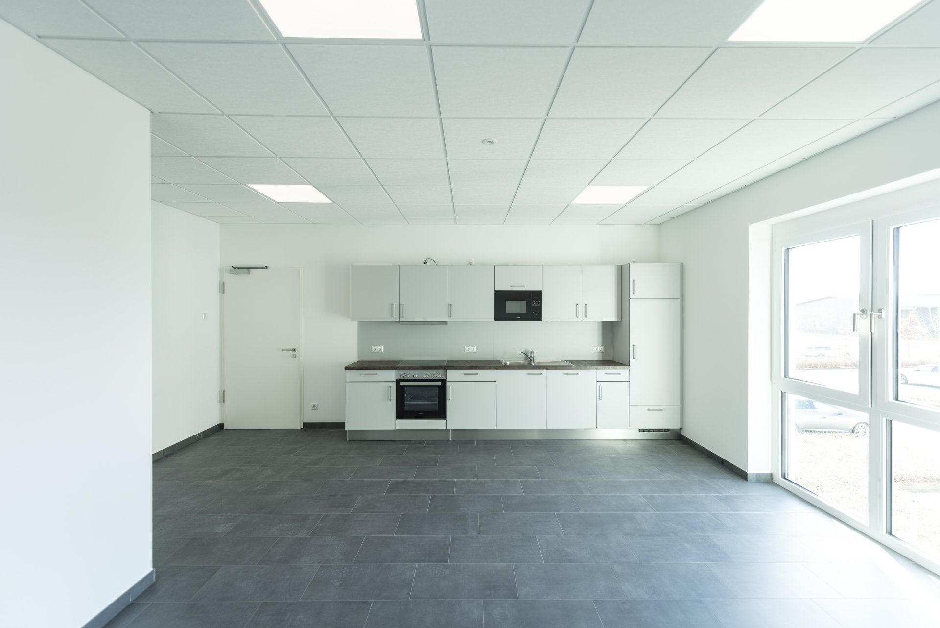 Leere, moderne Küche mit weißen Schränken, dunklem Boden und großen Fenstern, die Tageslicht hereinlassen