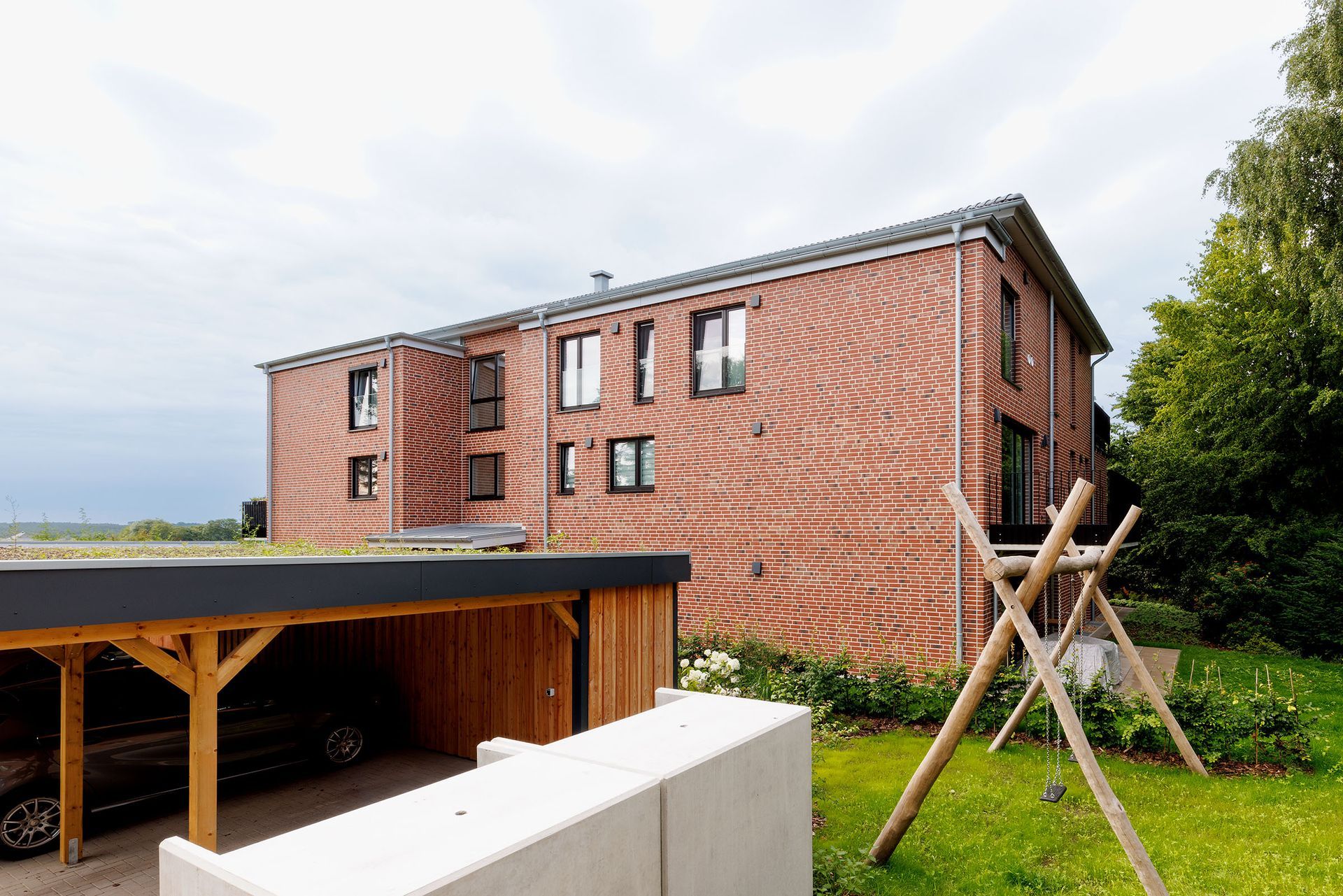 Rotes Backsteinhaus mit hölzernem Carport, grünem Rasen und Stützpfählen in ländlicher Umgebung