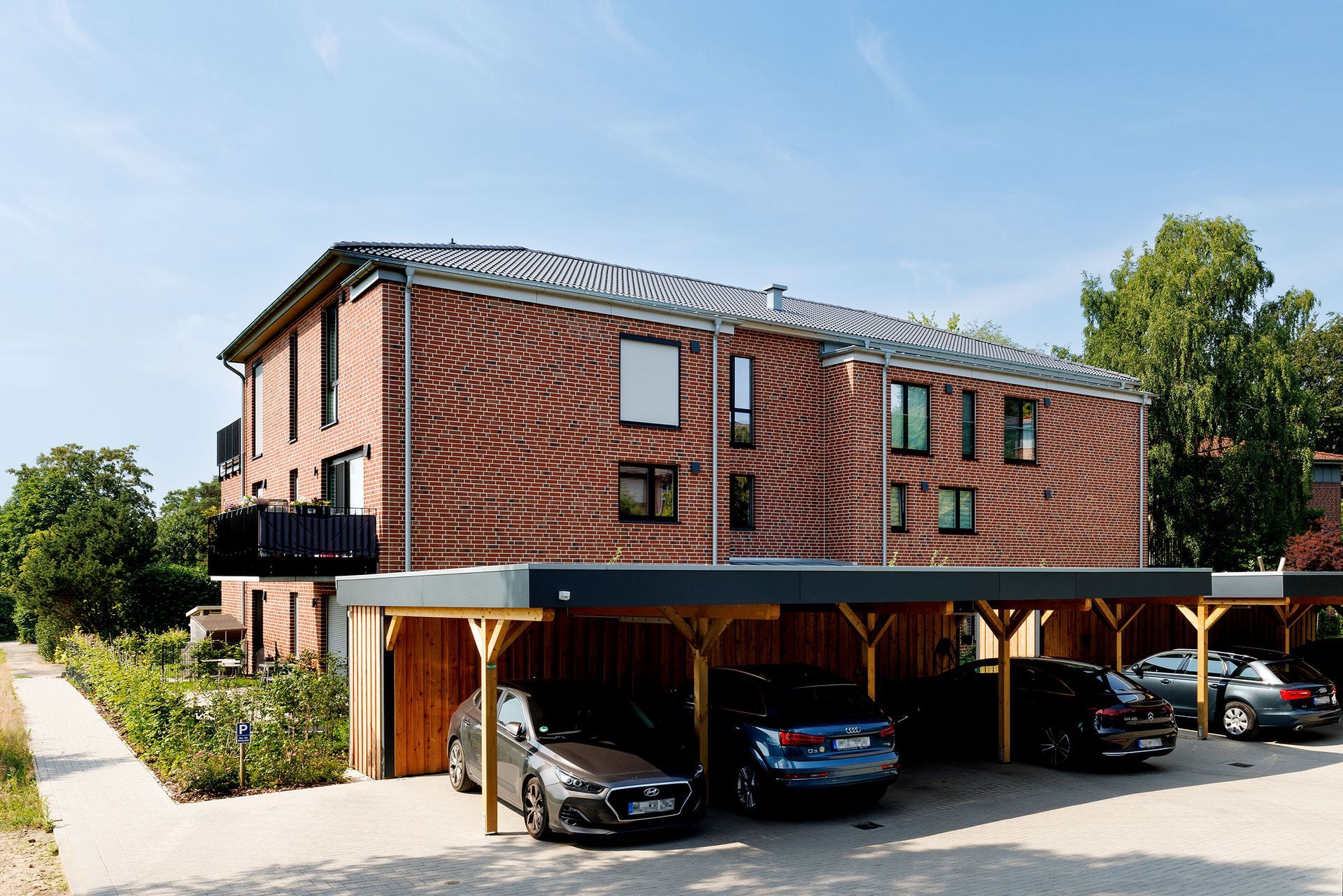 Backstein-Mehrfamilienhaus mit überdachtem Parkplatz und Autos, von der Straße aus an einem sonnigen Tag betrachtet