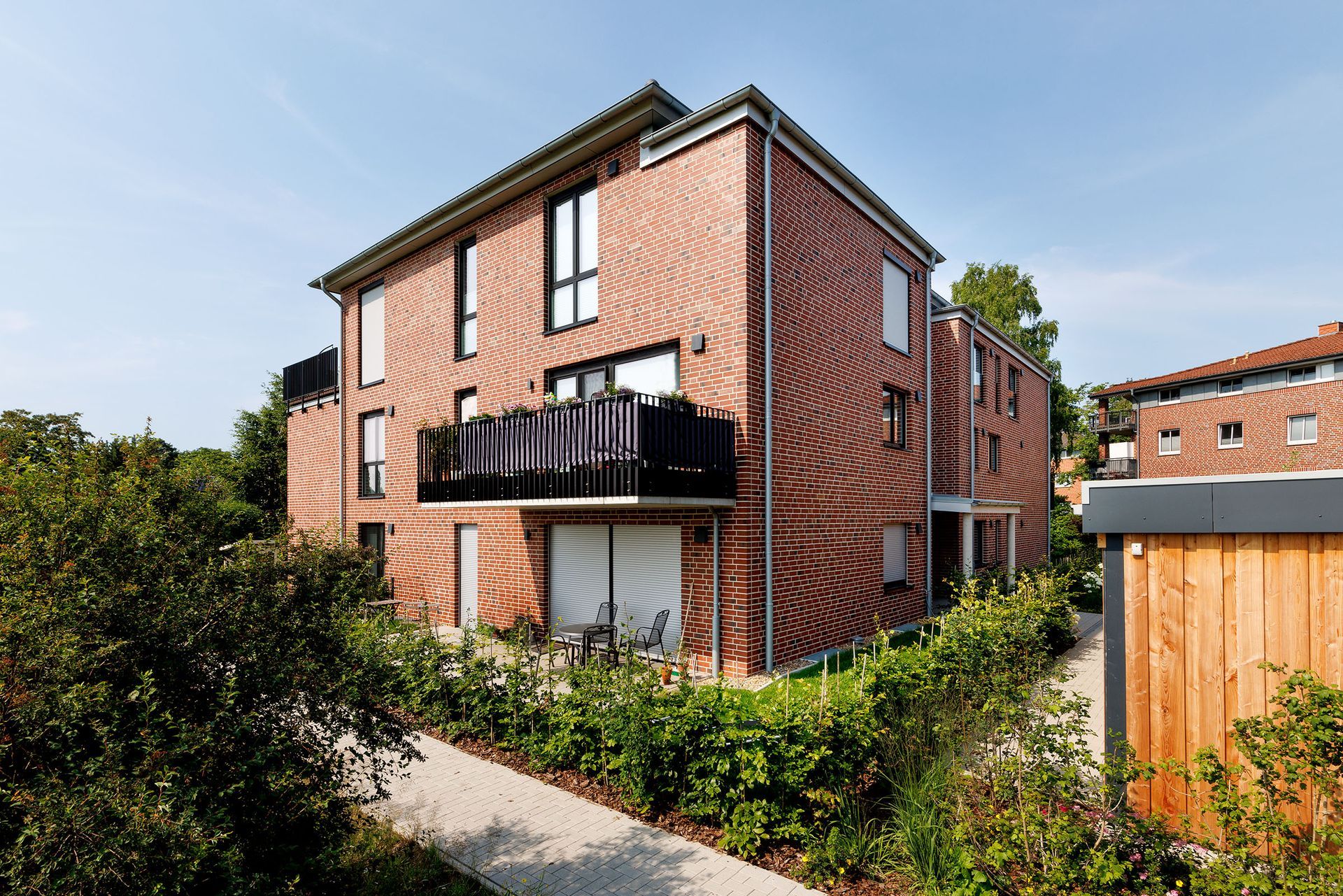 Reihenhaus aus rotem Backstein mit schwarzem Balkon und Fenstern, gesehen von einem Gartenweg neben dichtem Grün.