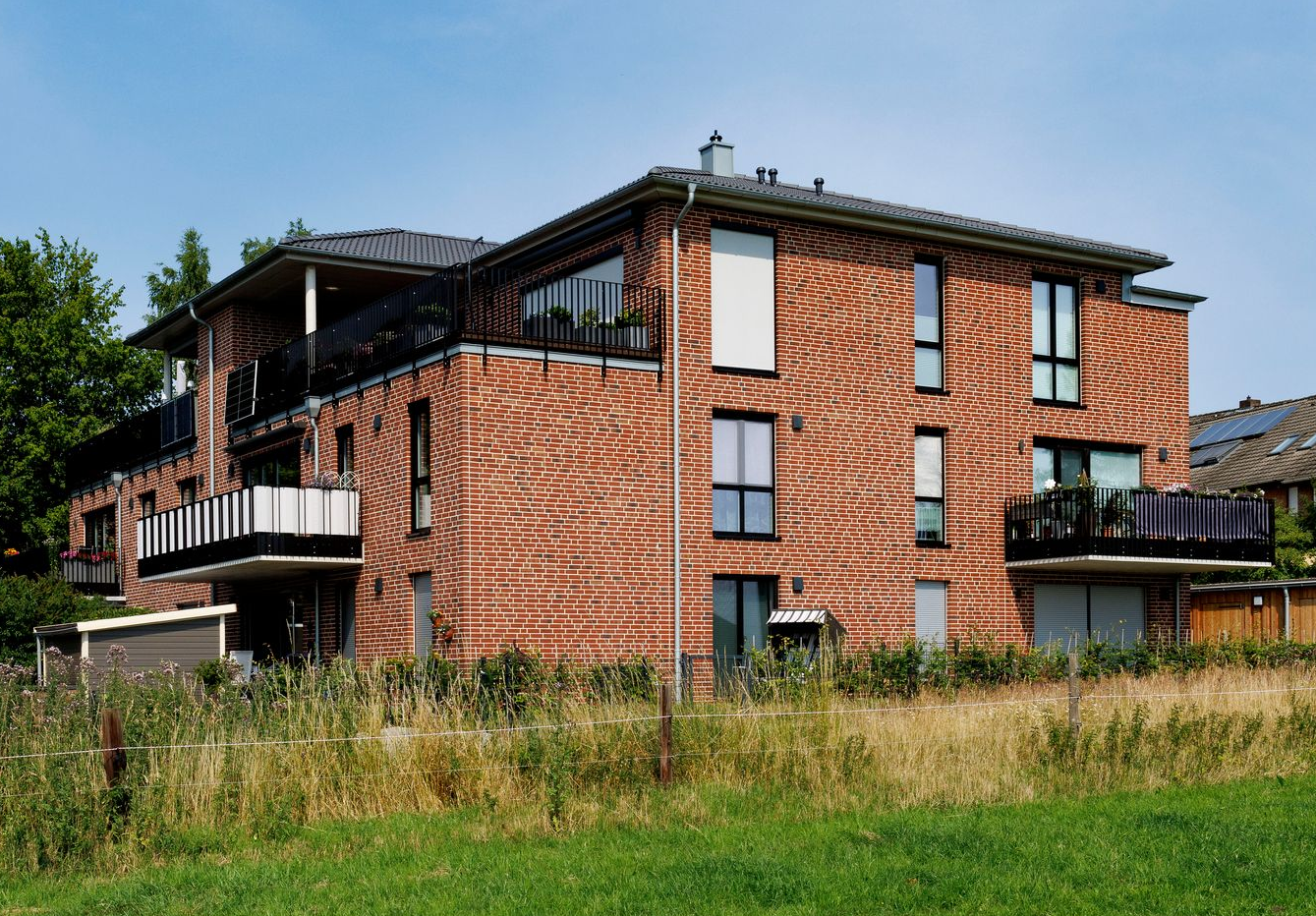 Rotes Backstein-Wohnhaus mit Balkonen und grasbewachsenem Vordergrund unter blauem Himmel