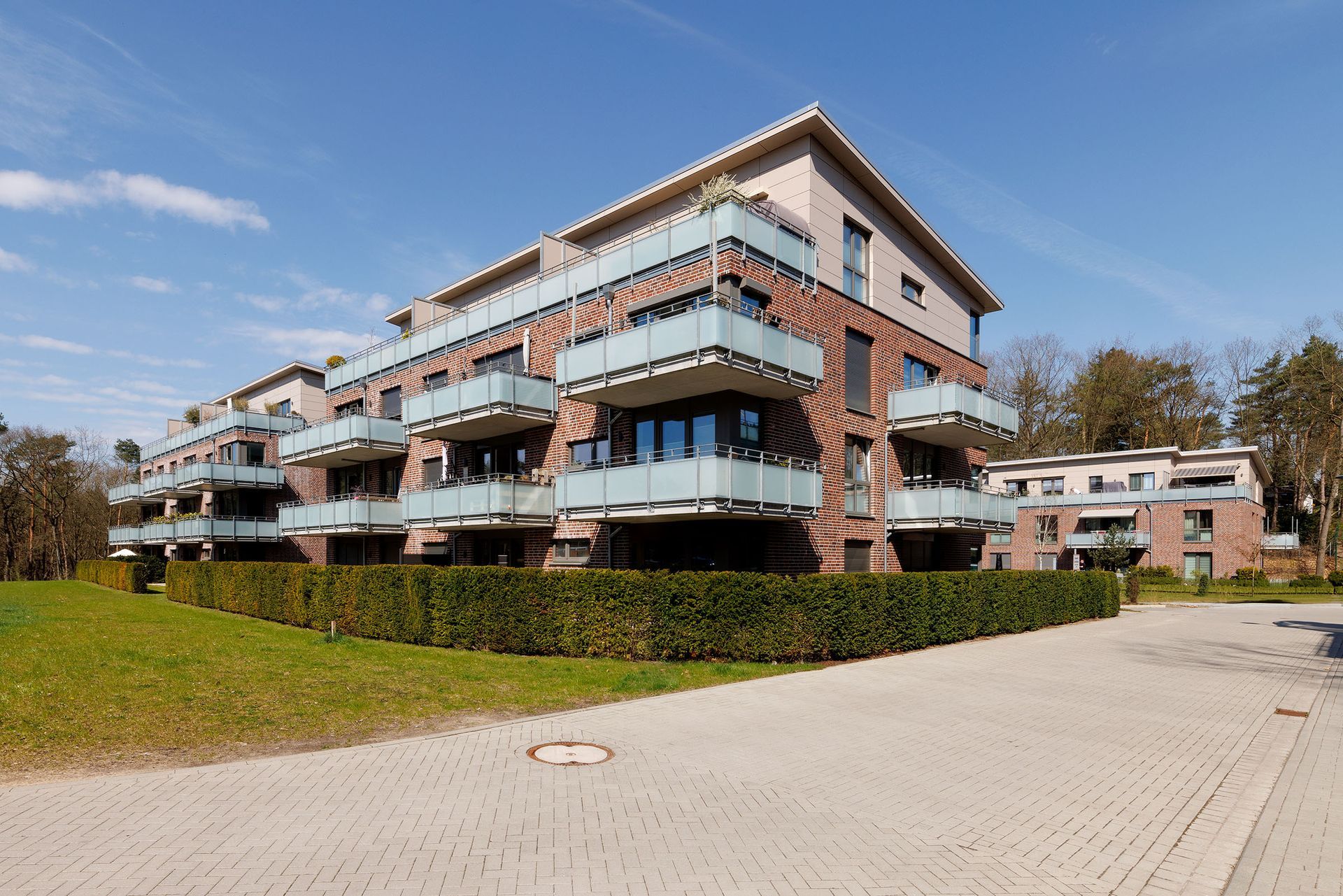 Modernes Wohnhaus mit Balkonen neben einem gepflasterten Weg und einem grasbewachsenen Hang unter blauem Himmel