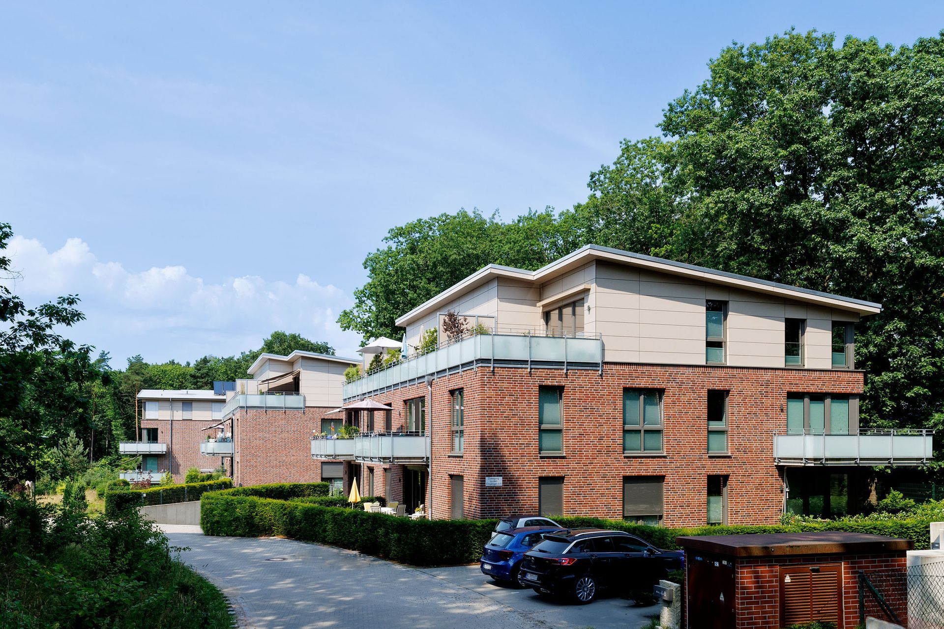 Ein Backsteinwohnhaus mit grünen Fenstern und geparkten Autos neben einer von Bäumen gesäumten Straße unter blauem Himmel