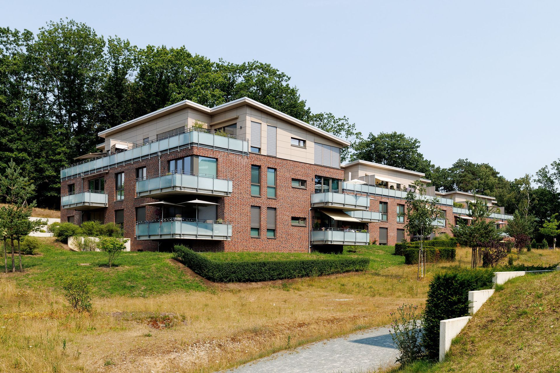 Ein Backstein-Mehrfamilienhaus mit Balkonen liegt an einem grasbewachsenen Hang und ist von Bäumen umgeben.