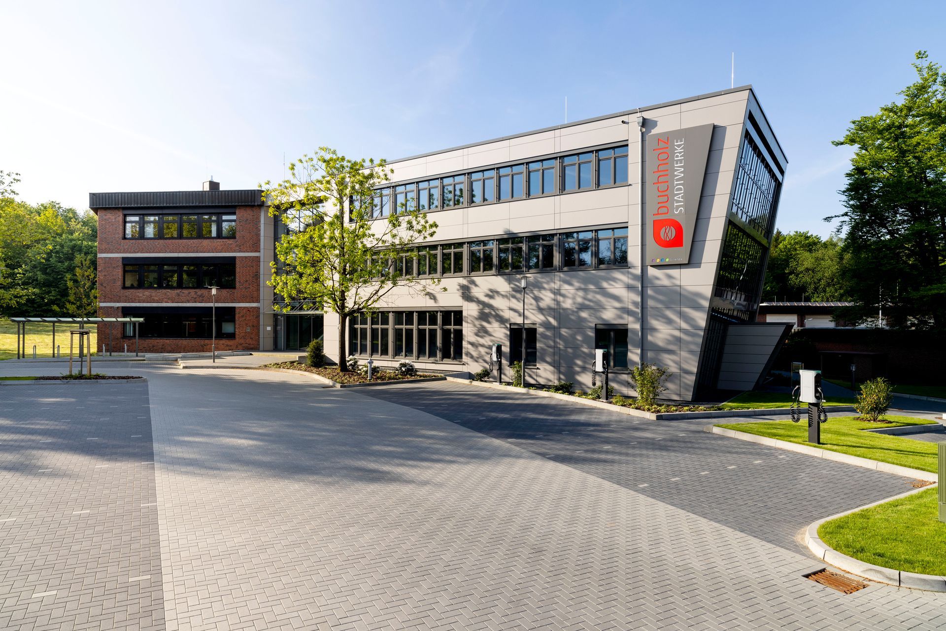 Modernes Bürogebäude mit Glasfassade, Backsteinflügel und gepflasterter Zufahrt unter klarem Himmel