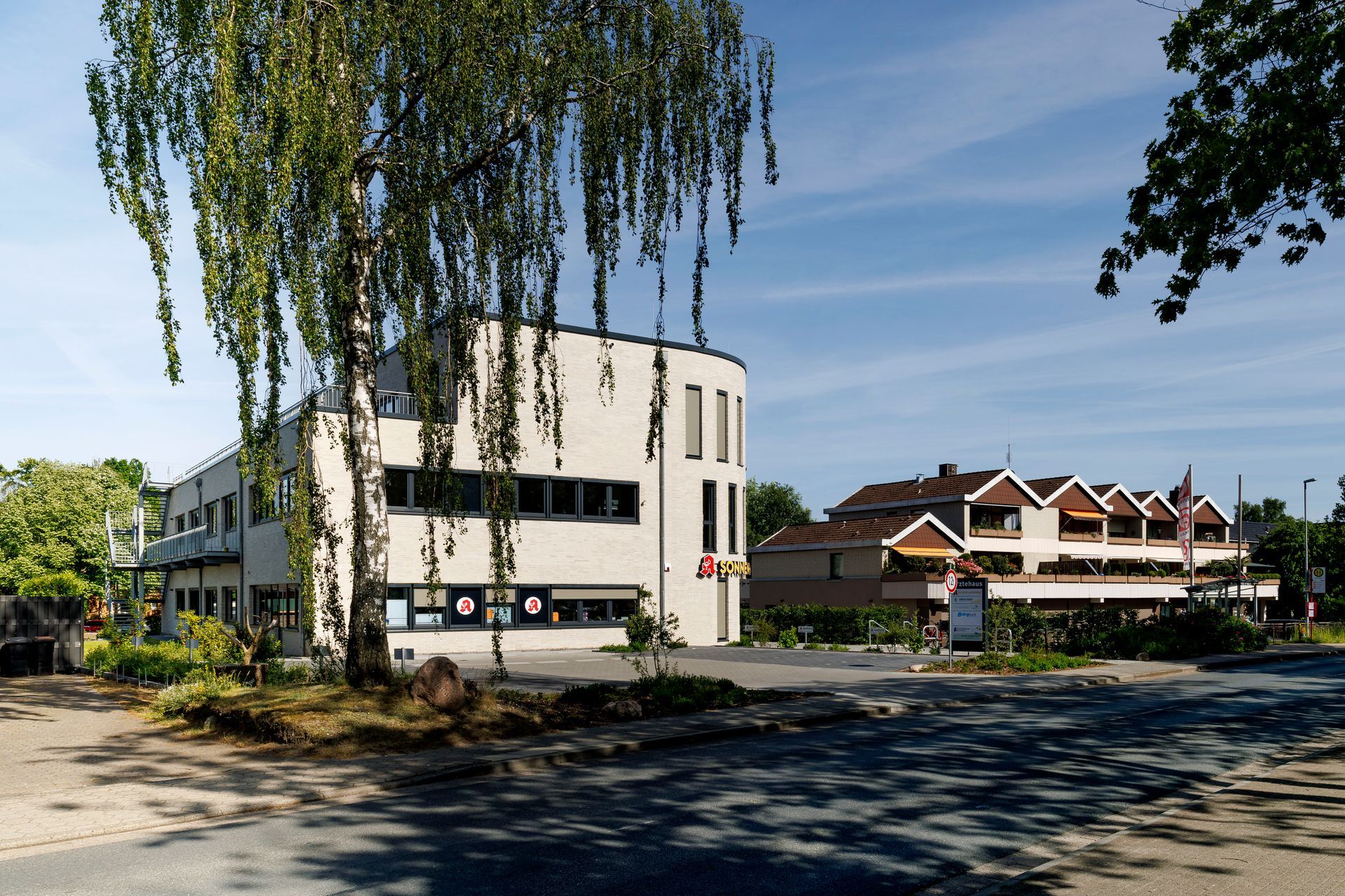 Modernes weißes Gebäude neben einer Straße, zwischen Bäumen und Häusern unter klarem blauen Himmel