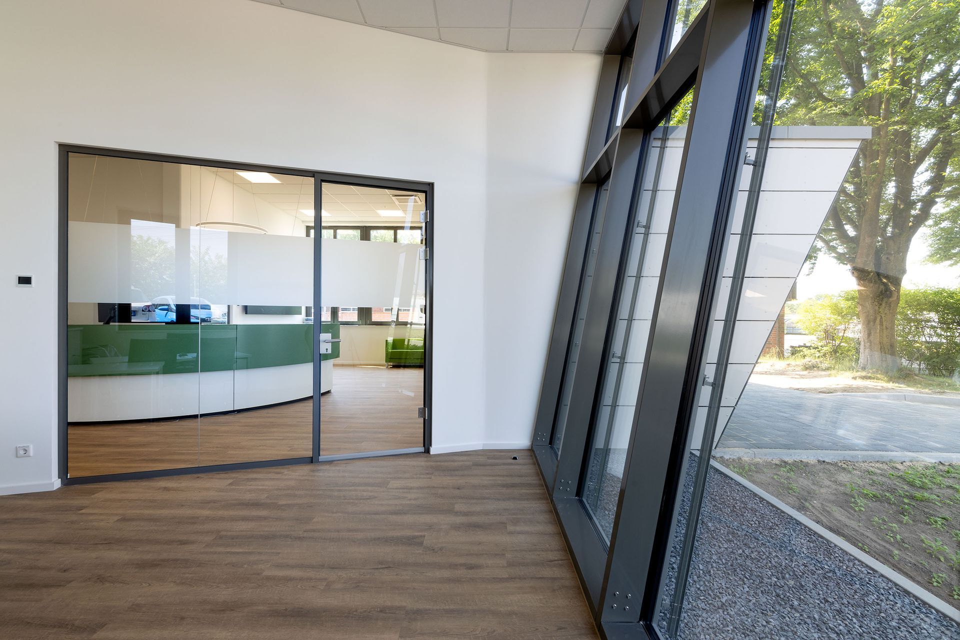 Leere Bürolobby mit Holzboden, Glastrennwand und großen Fenstern mit Blick auf Bäume.