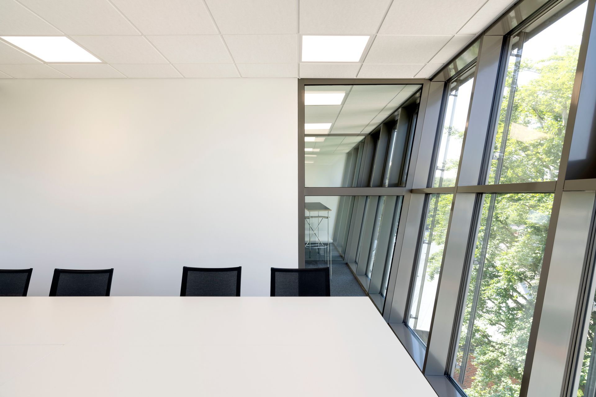 Heller, moderner Konferenzraum mit weißem Tisch, schwarzen Stühlen und großen Fenstern mit Blick auf die Bäume