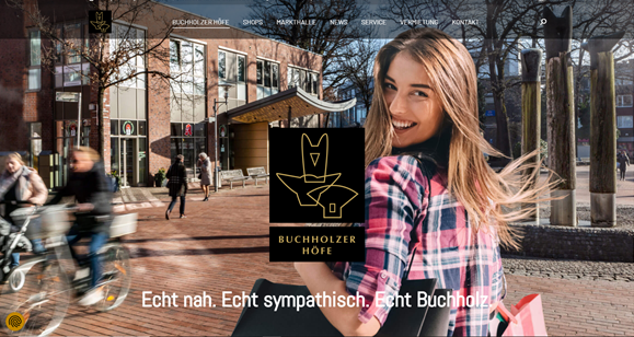 Eine Frau lächelt auf einem Campusweg und hält ein schwarzes Schild mit einem gezeichneten Katzenlogo in der Hand.