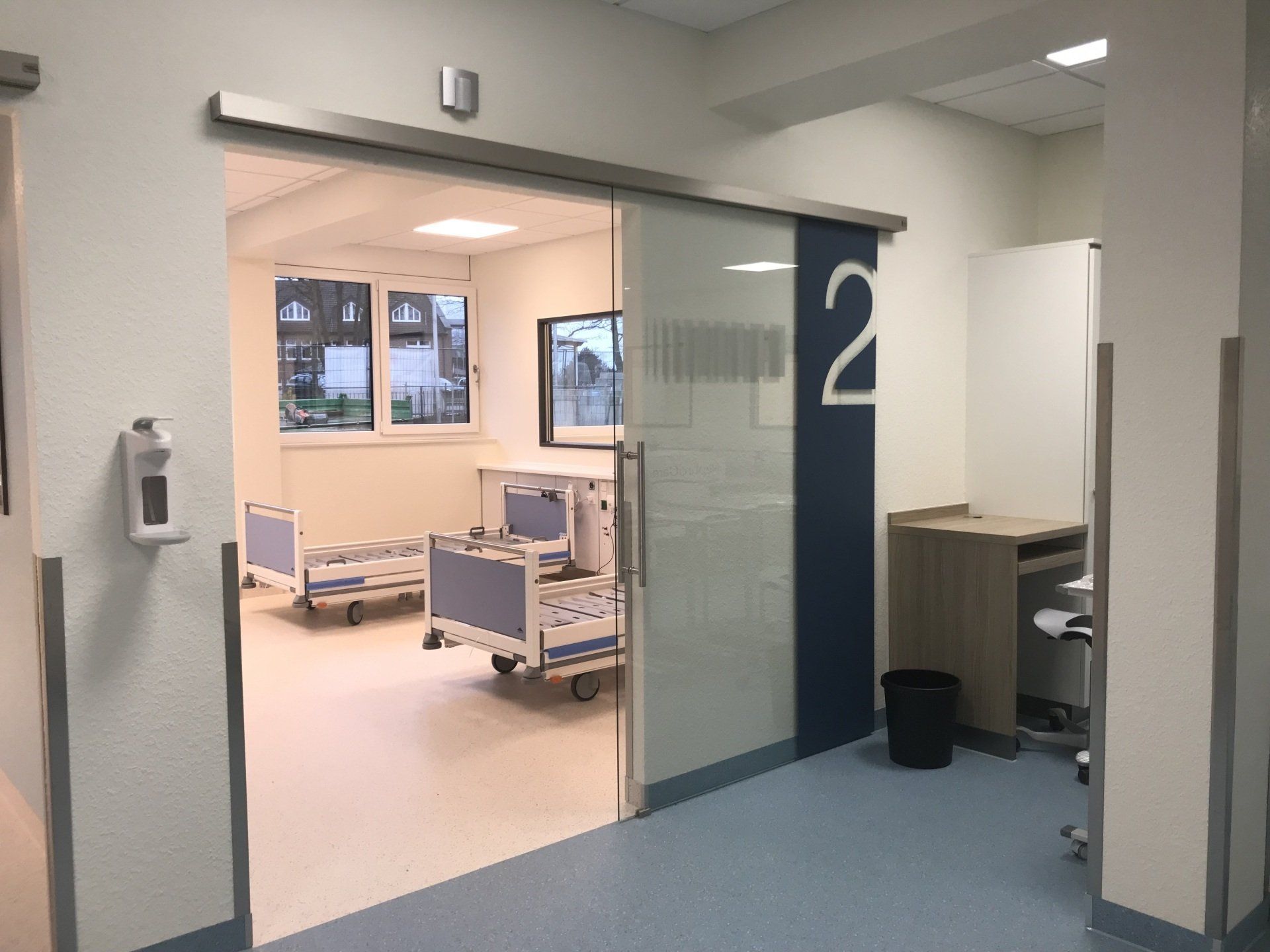 Krankenhausflur mit offenem Zimmereingang, blauem Bodenbelag und Betten, die in Zimmer 2 sichtbar sind.