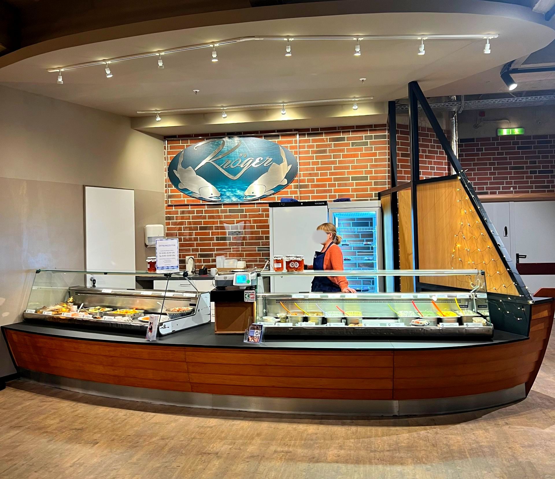 Bäckerei- oder Feinkosttheke mit Gebäck, Backsteinwand und einem Mitarbeiter hinter der Auslage