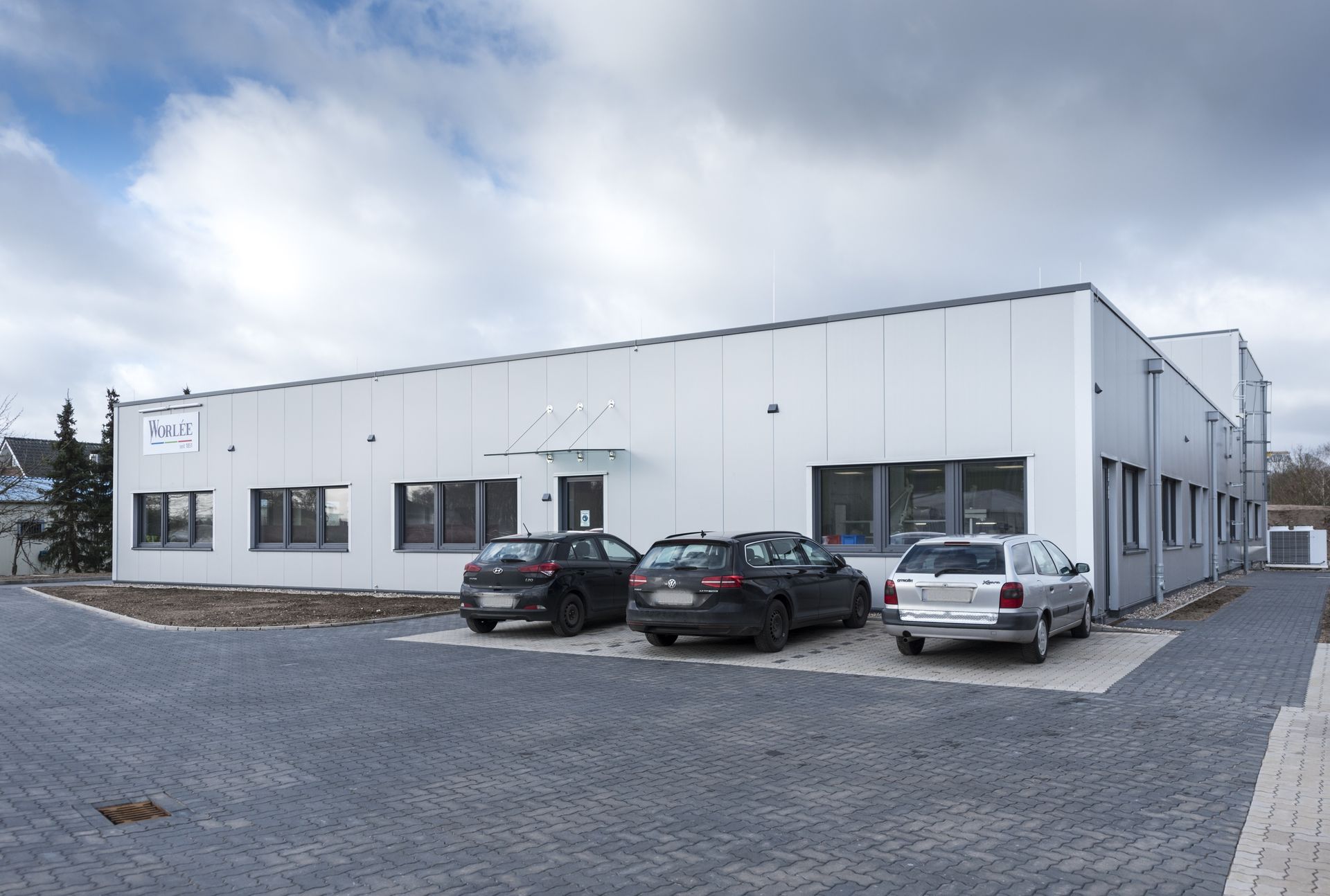 Modernes weißes Industriegebäude mit drei geparkten Autos auf einem gepflasterten Parkplatz unter bewölktem Himmel