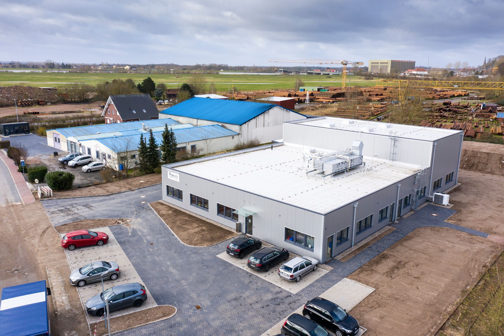 Luftaufnahme eines modernen Industriegebäudes mit blauen Dächern, geparkten Autos und umliegenden Feldern