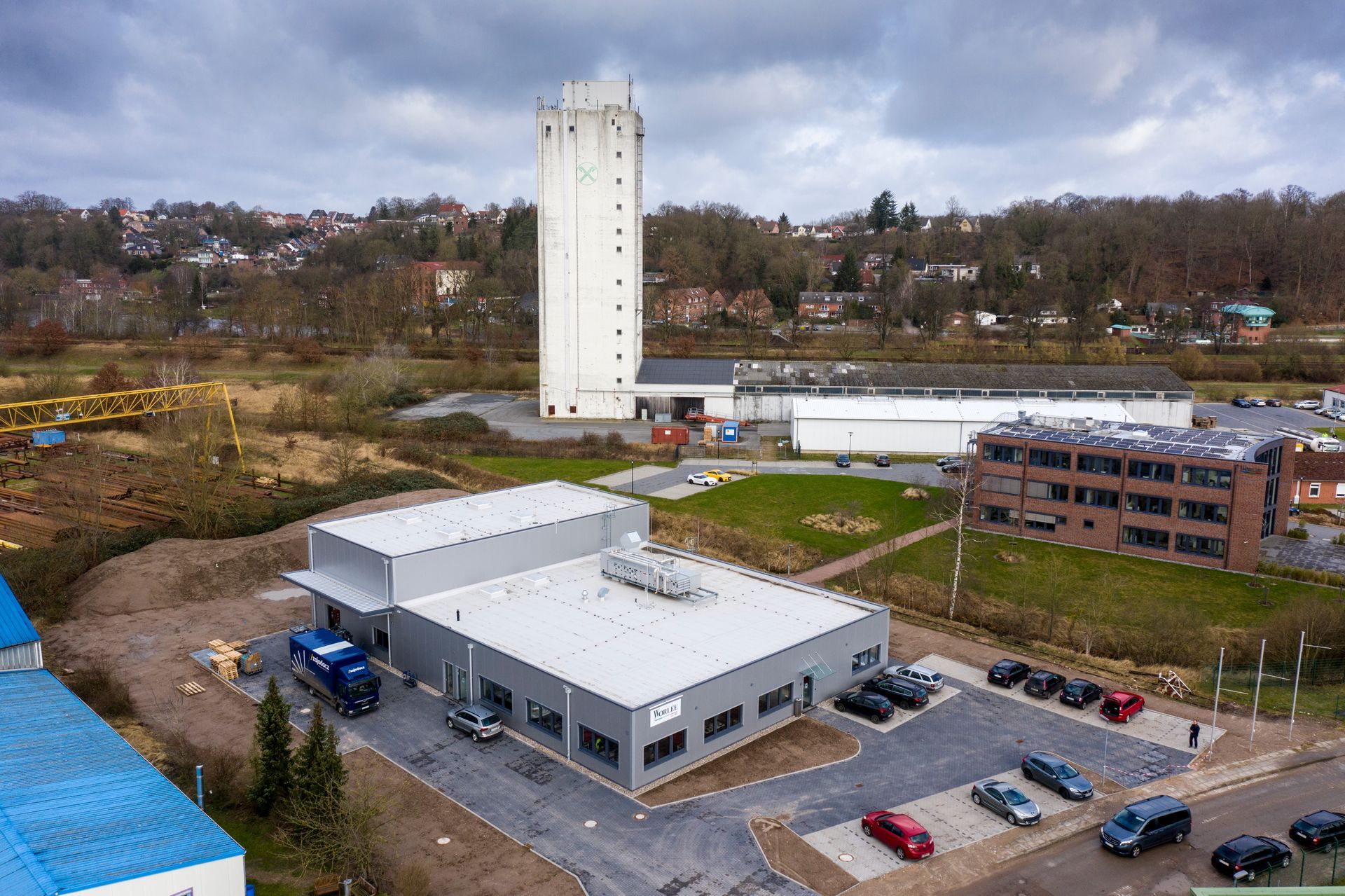 Luftaufnahme eines Industriegeländes mit einem hohen weißen Turm, grauen Gebäuden und geparkten Autos