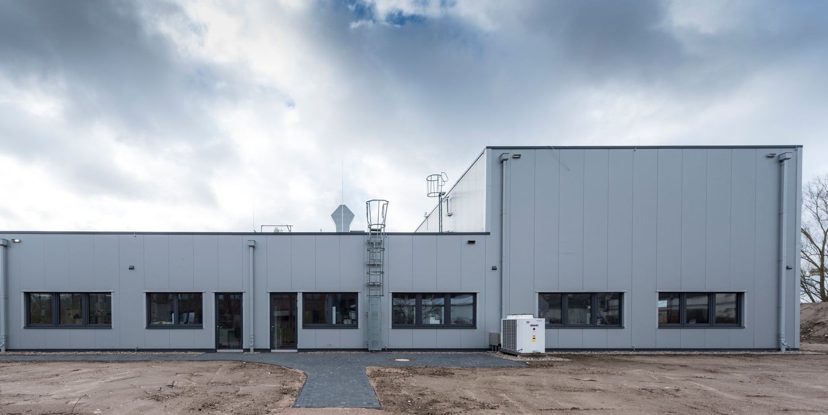 Niedriges Industriegebäude mit grauer Fassade und Fensterreihen unter einem bewölkten Himmel