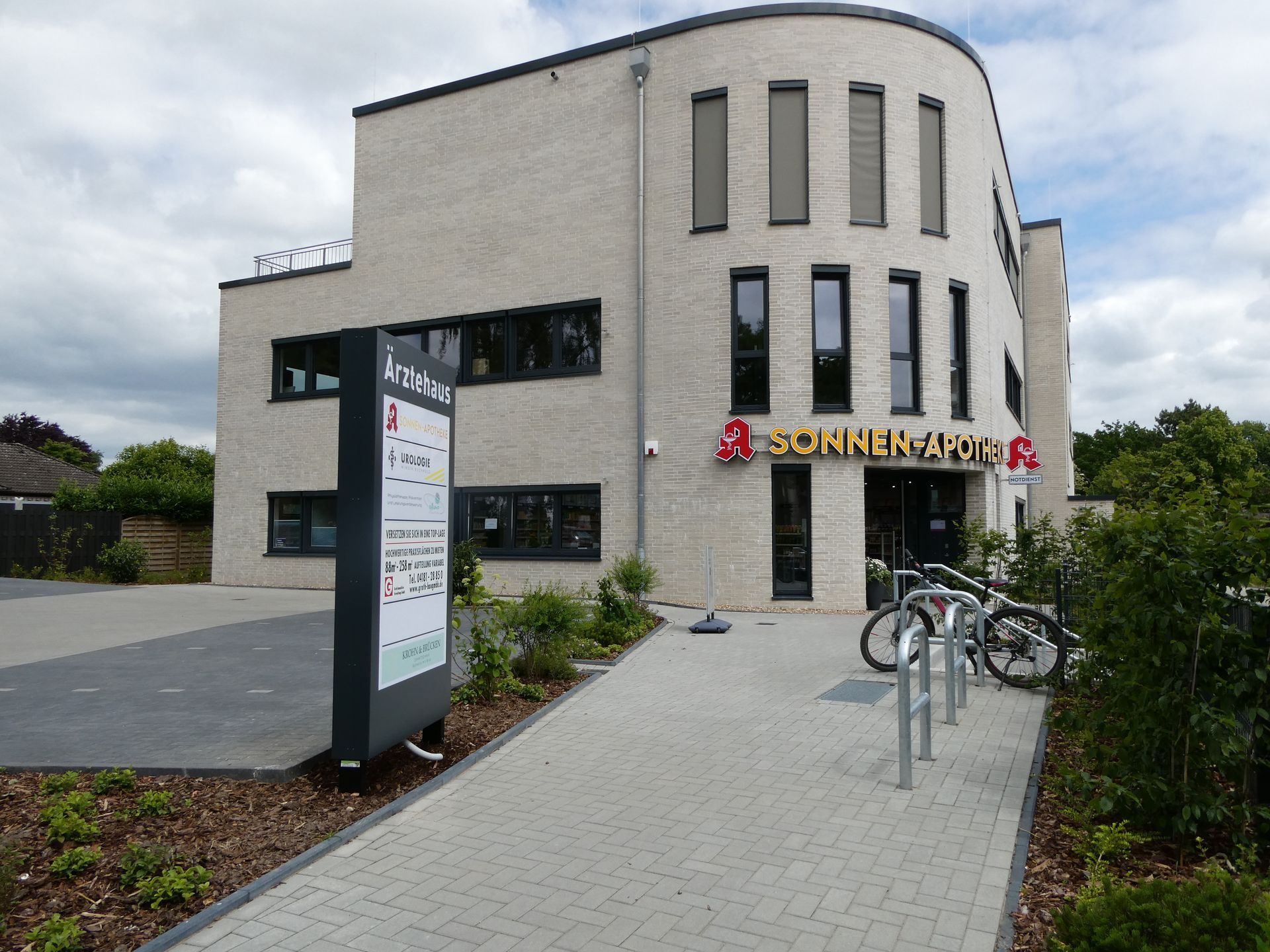 Modernes beigefarbenes Bürogebäude mit Schild, gepflastertem Gehweg und Fahrrädern davor.
