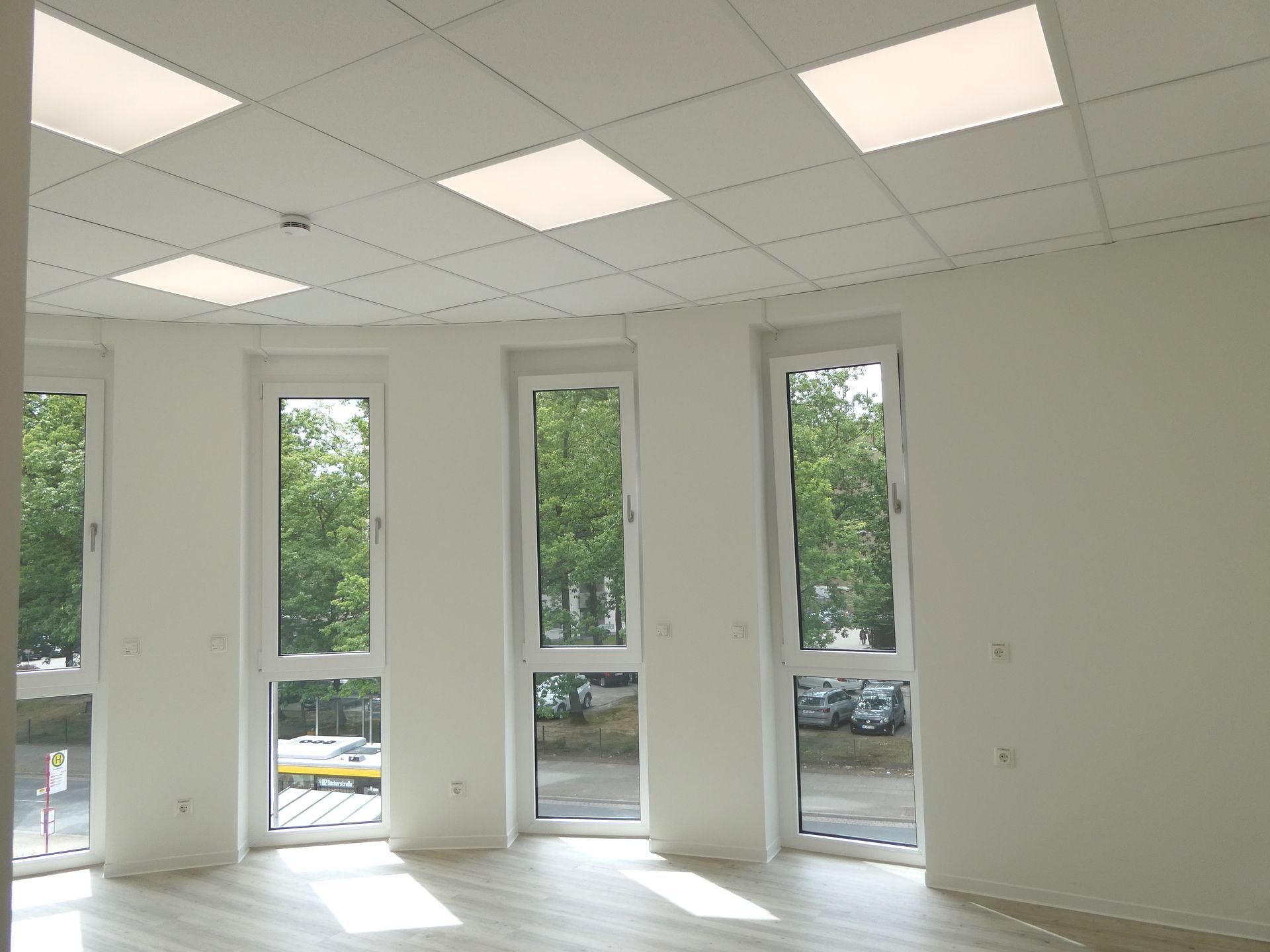 Leerer weißer Raum mit großen Fenstern und Deckenleuchten, mit Blick auf die Bäume draußen