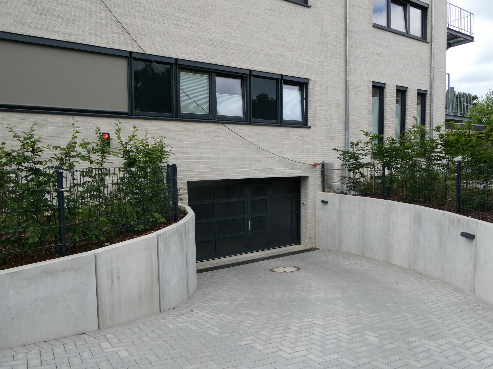 Eine Betonauffahrt führt zu einem modernen Gebäude mit Garagentoren im Untergeschoss und angelegten Pflanzbeeten.