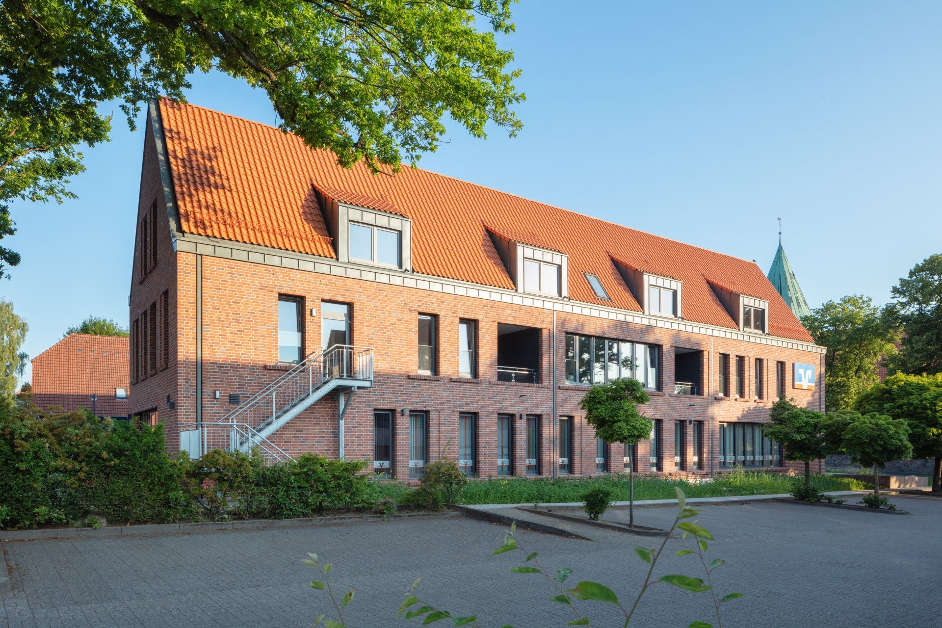 Backstein-Mehrfamilienhaus mit rotem Ziegeldach und Bäumen neben einem gepflasterten Grundstück