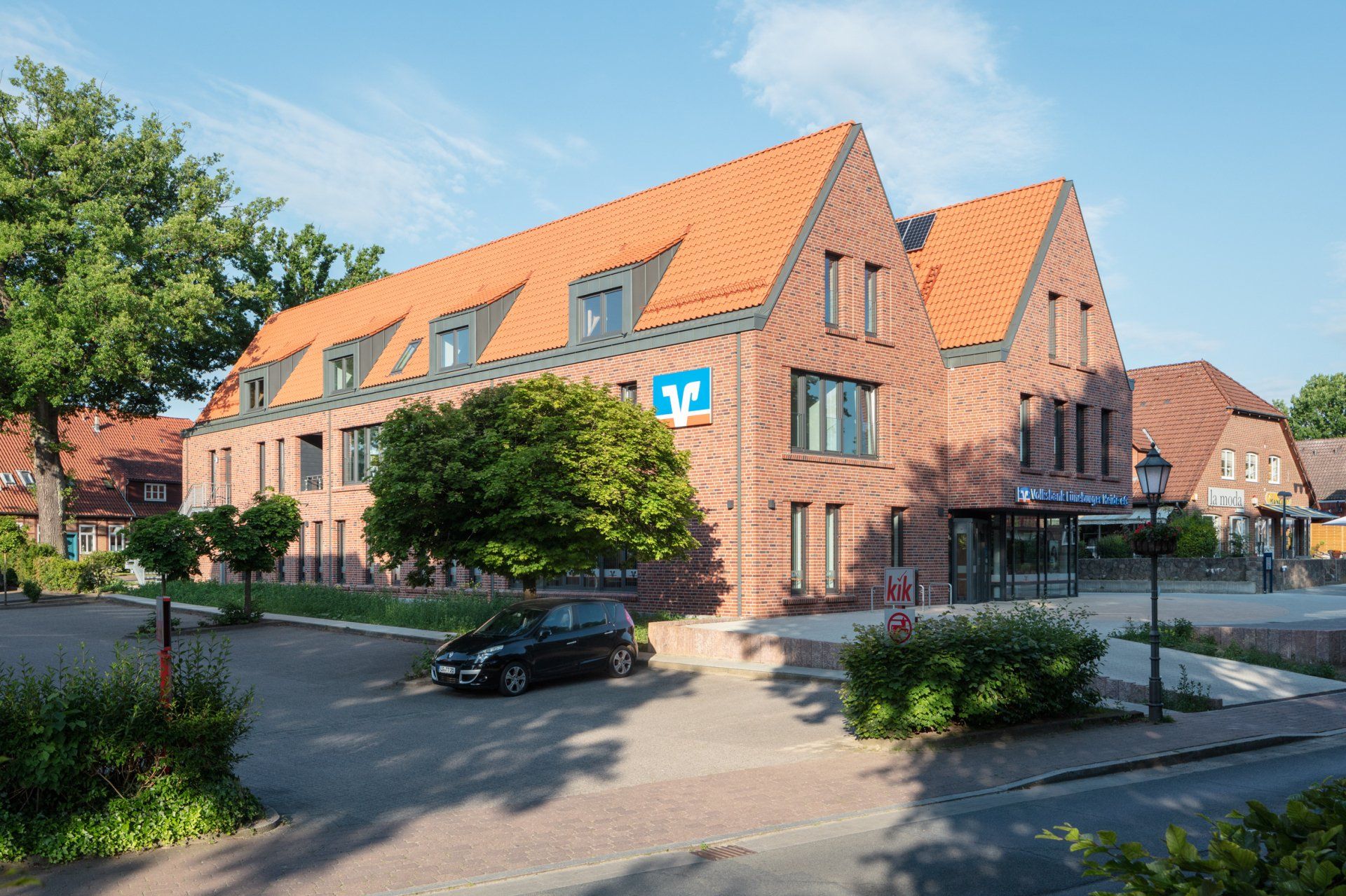 Backsteingebäude mit rotem Dach und Dachgauben an einer Straße, mit Bäumen und geparkten Autos