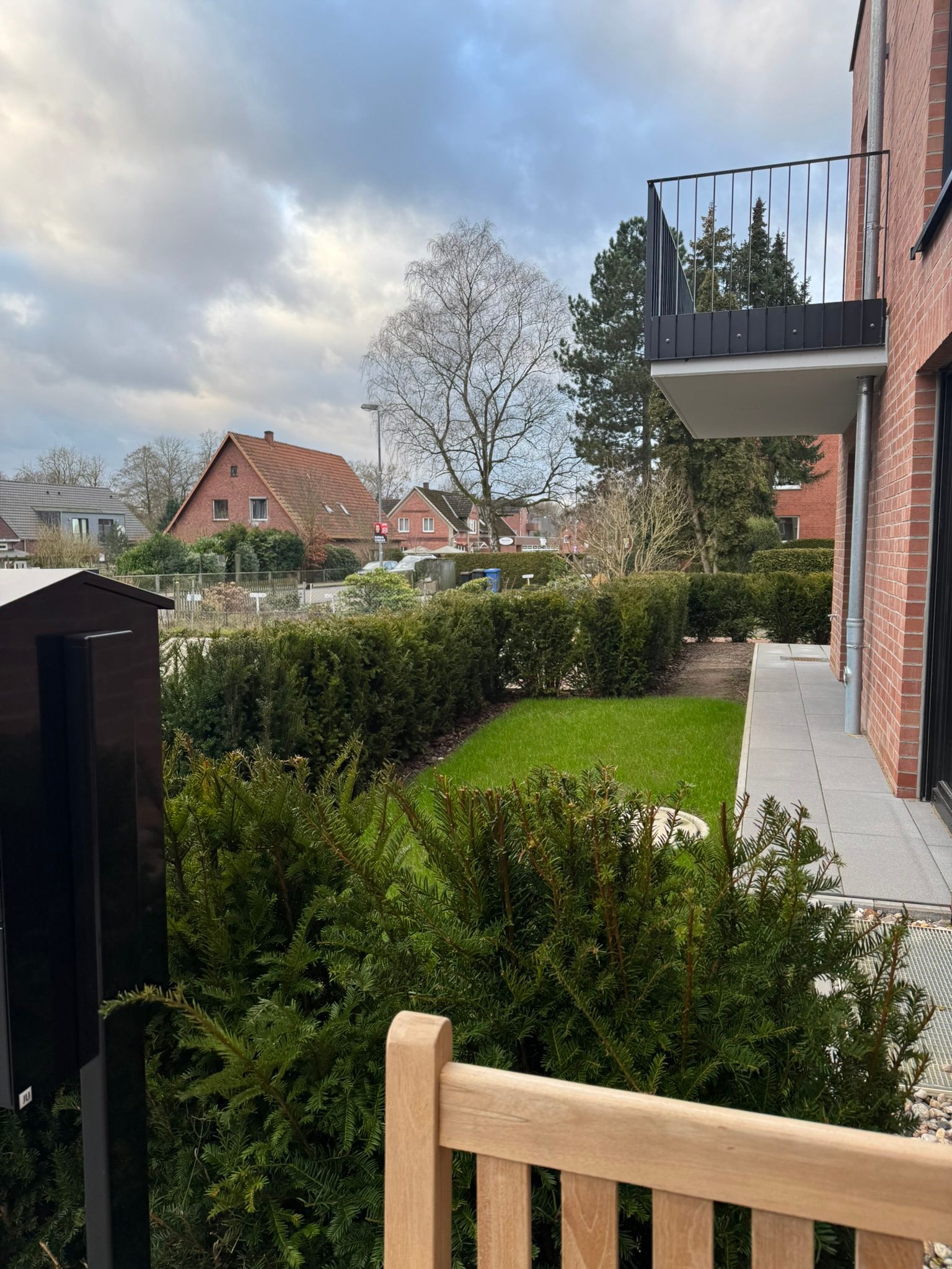 Hinterhofgarten neben einem Backsteinhaus mit Balkon, grünen Hecken und bewölktem Himmel.