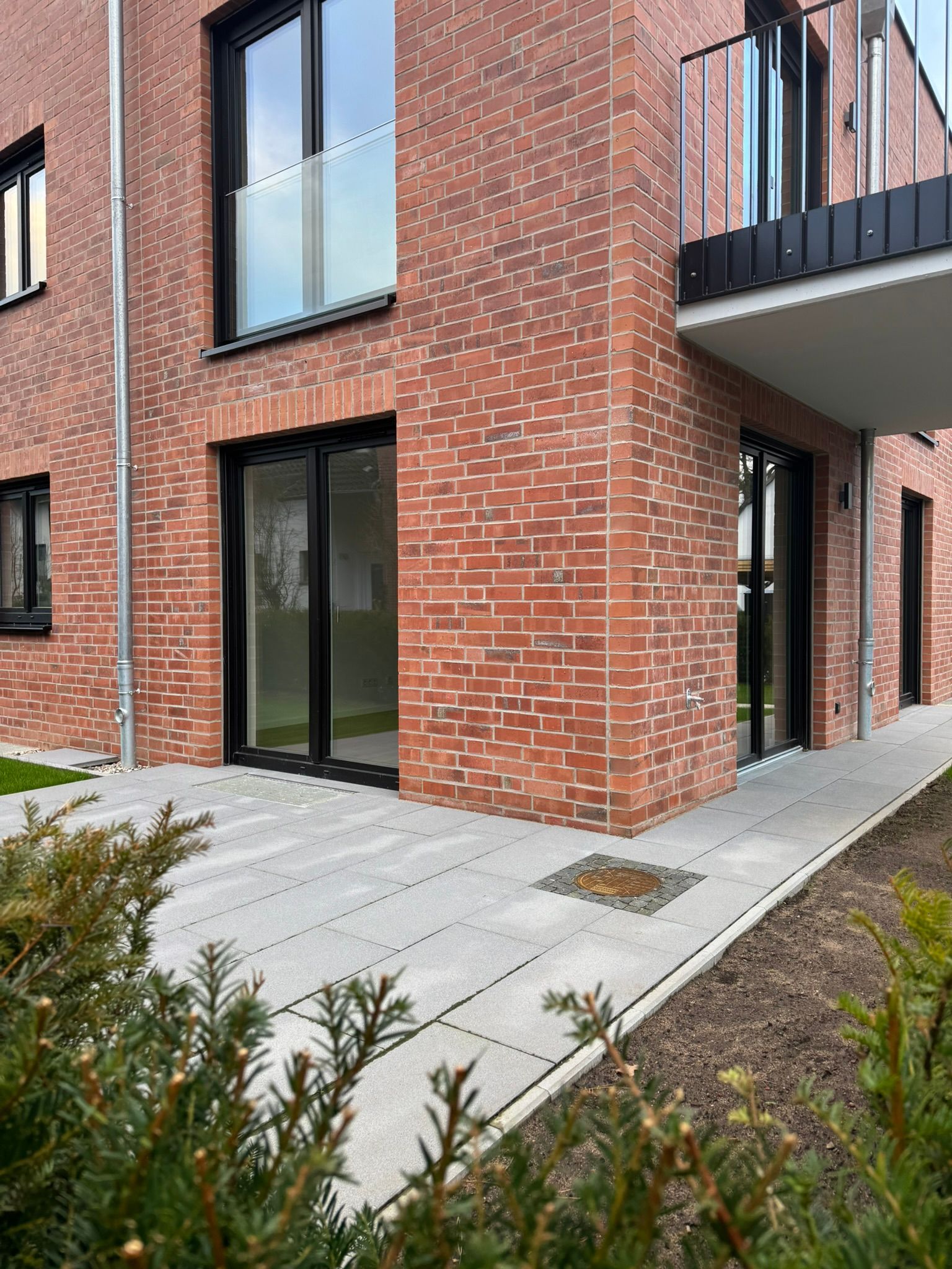 Mehrfamilienhaus aus Backstein mit Glastüren, Balkon und gepflastertem Terrassenweg neben einer Grünanlage.