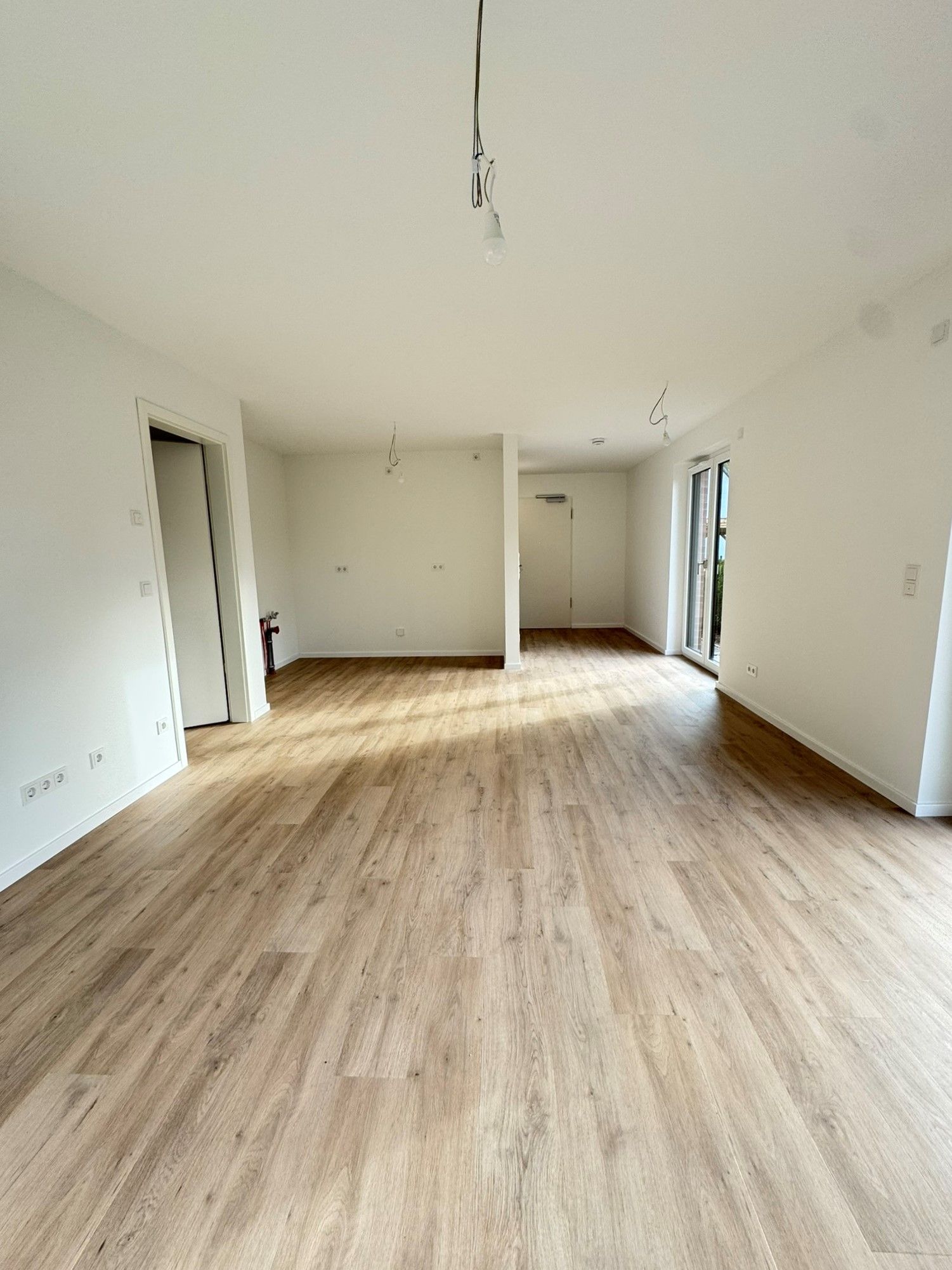 Ein leerer, weißer Raum mit hellem Holzboden und herabhängenden Deckenkabeln, Blickrichtung Fenster am anderen Ende
